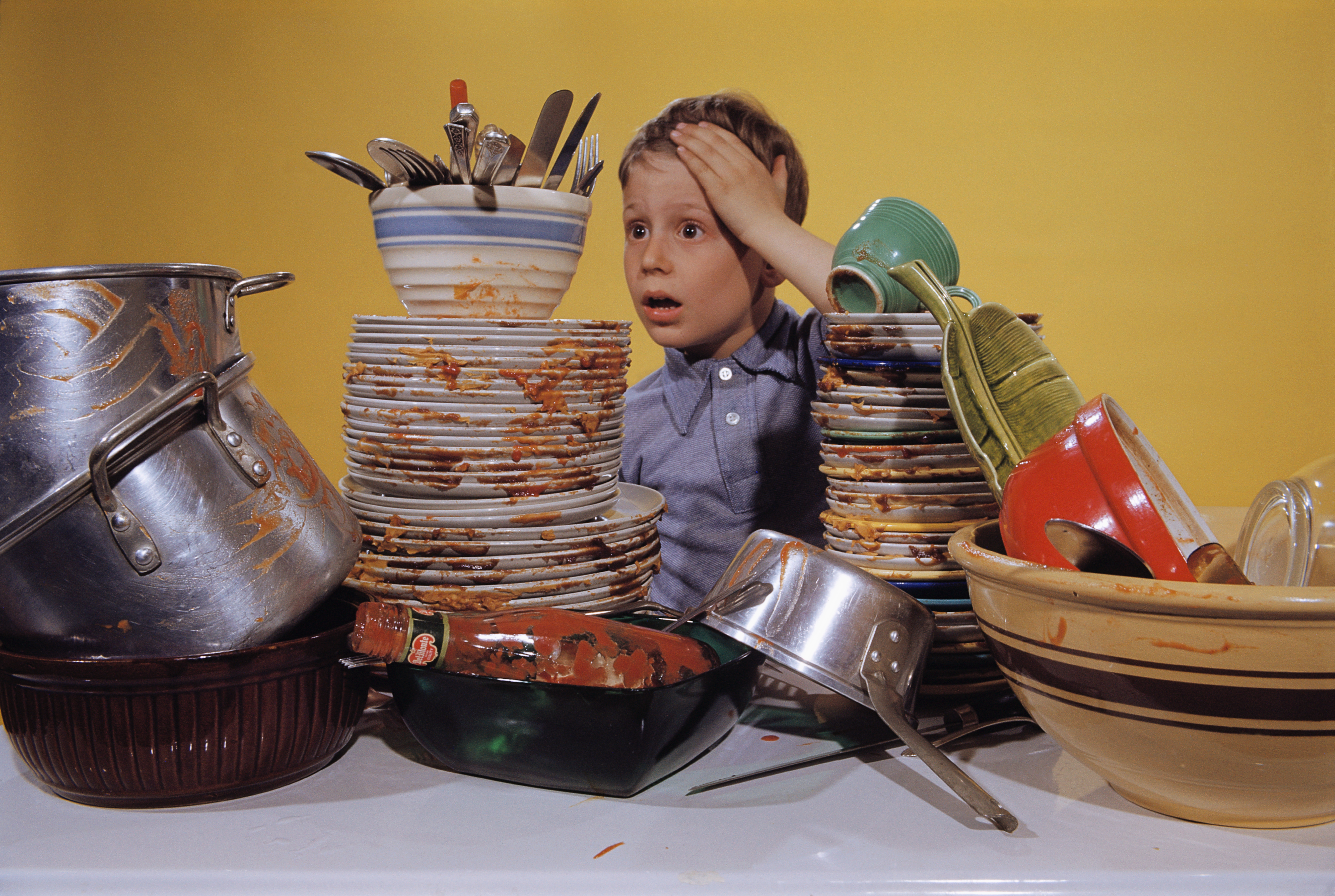 A young boy with an overwhelmed expression on his face stands behind a counter, surrounded by stacks of dirty dishes.