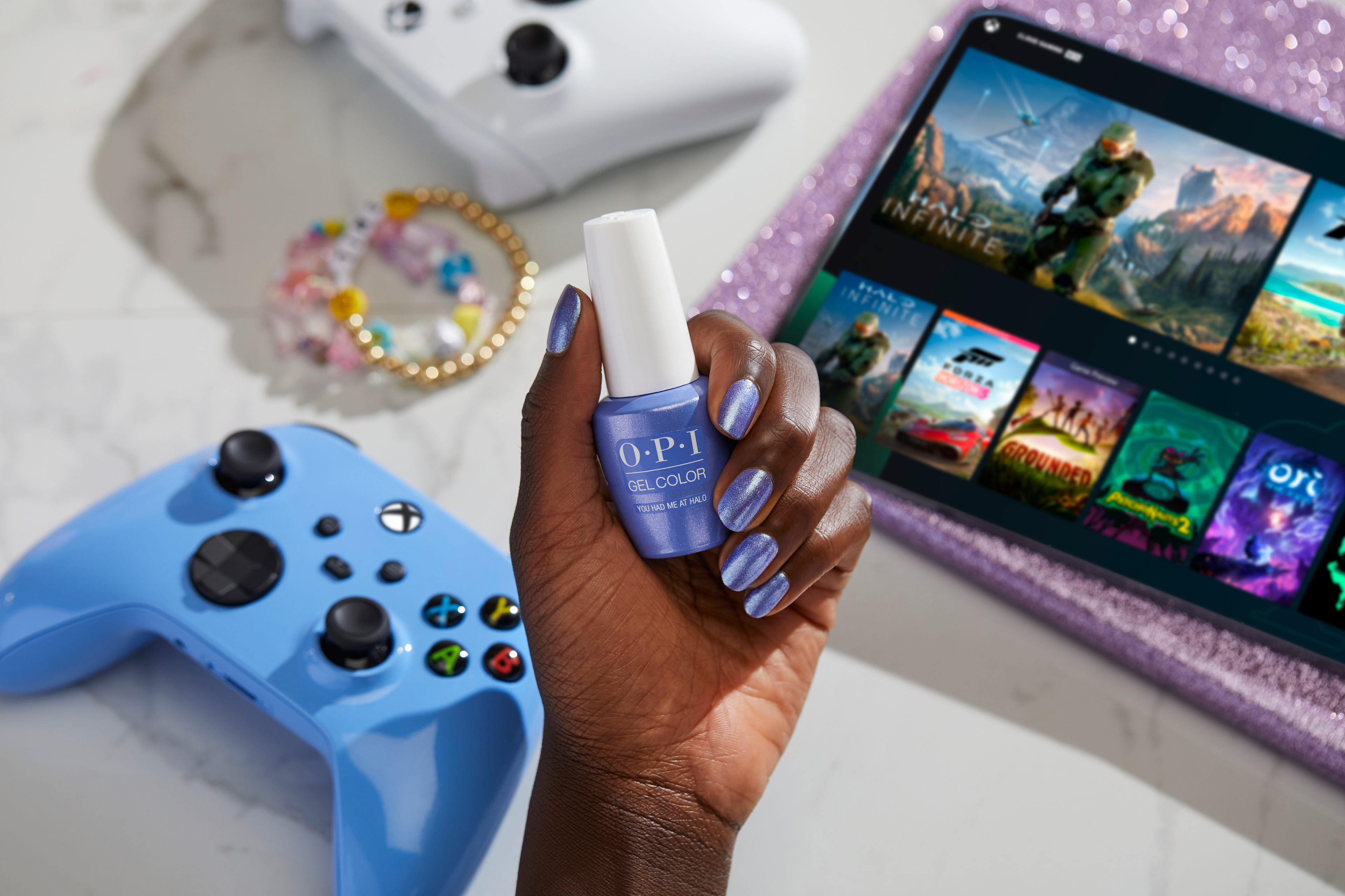a brown-skinned hand holding up a bottle of metallic periwinkle nail polish with a label saying “You Had Me at Halo,” with a blue Xbox controller in the background