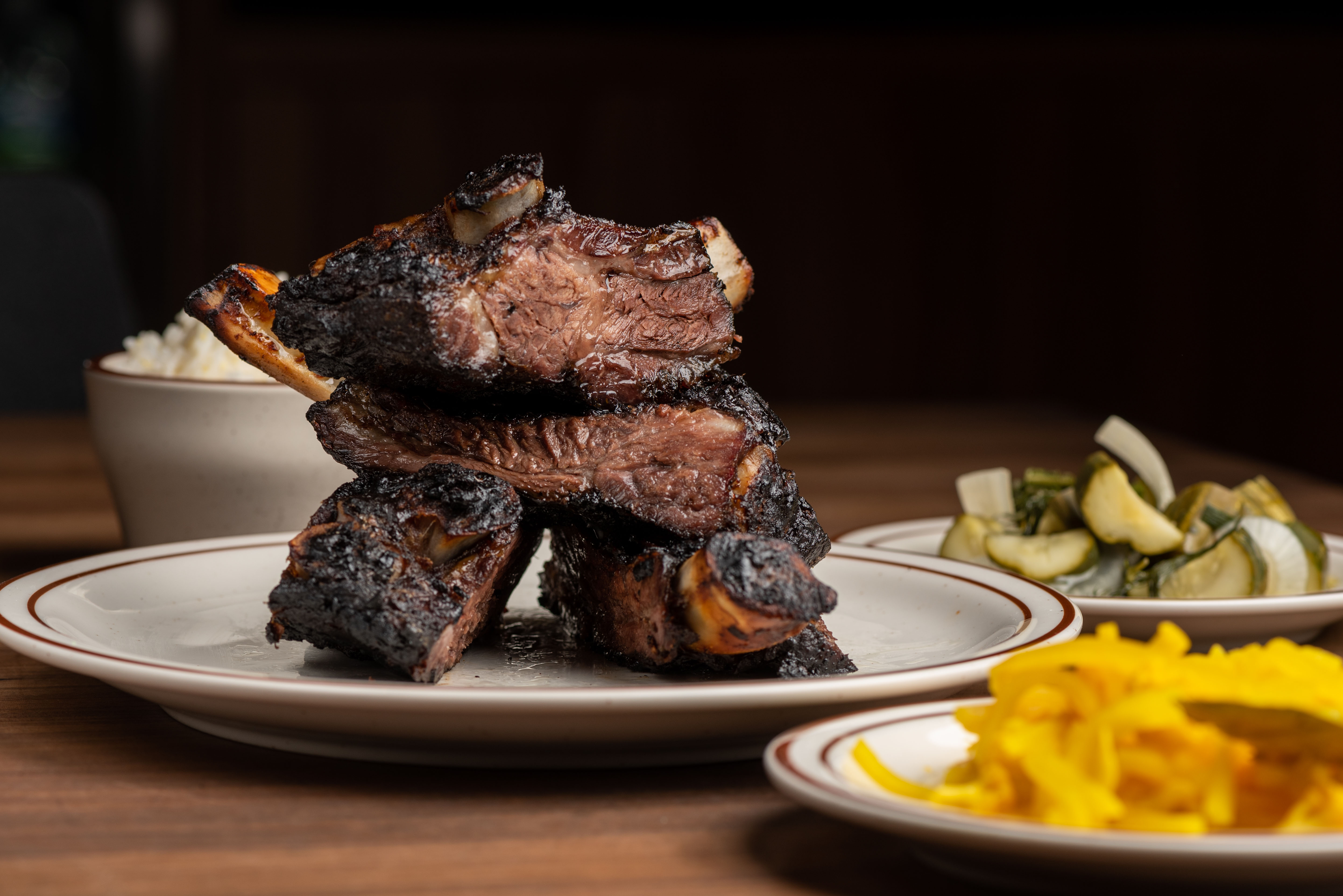 A side shot of charred ribs stacked onto each other on a white plate and wooden table.