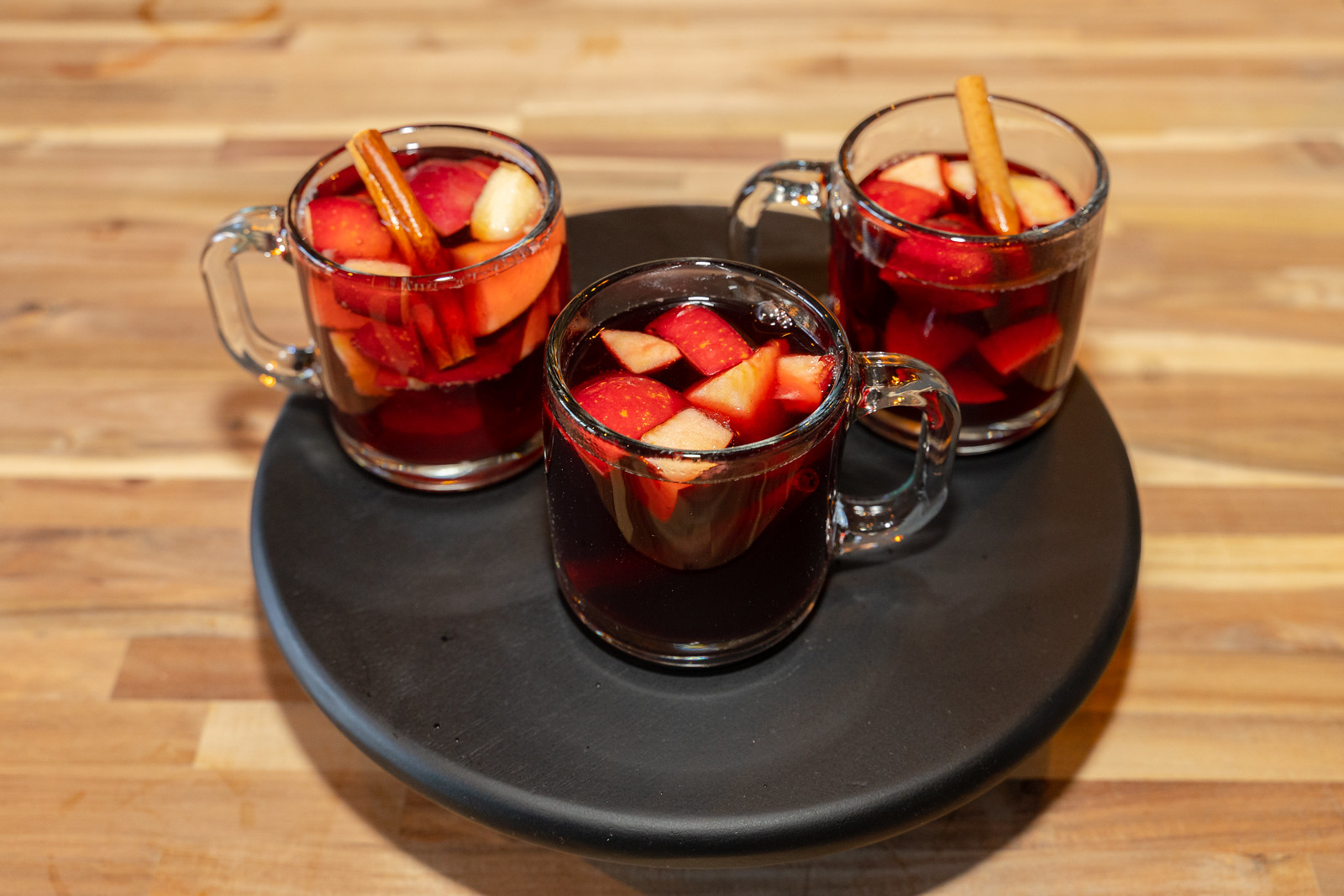 A black circular tray with three glass mugs with dark red drinks with cut fruit and cinnamon sticks. 