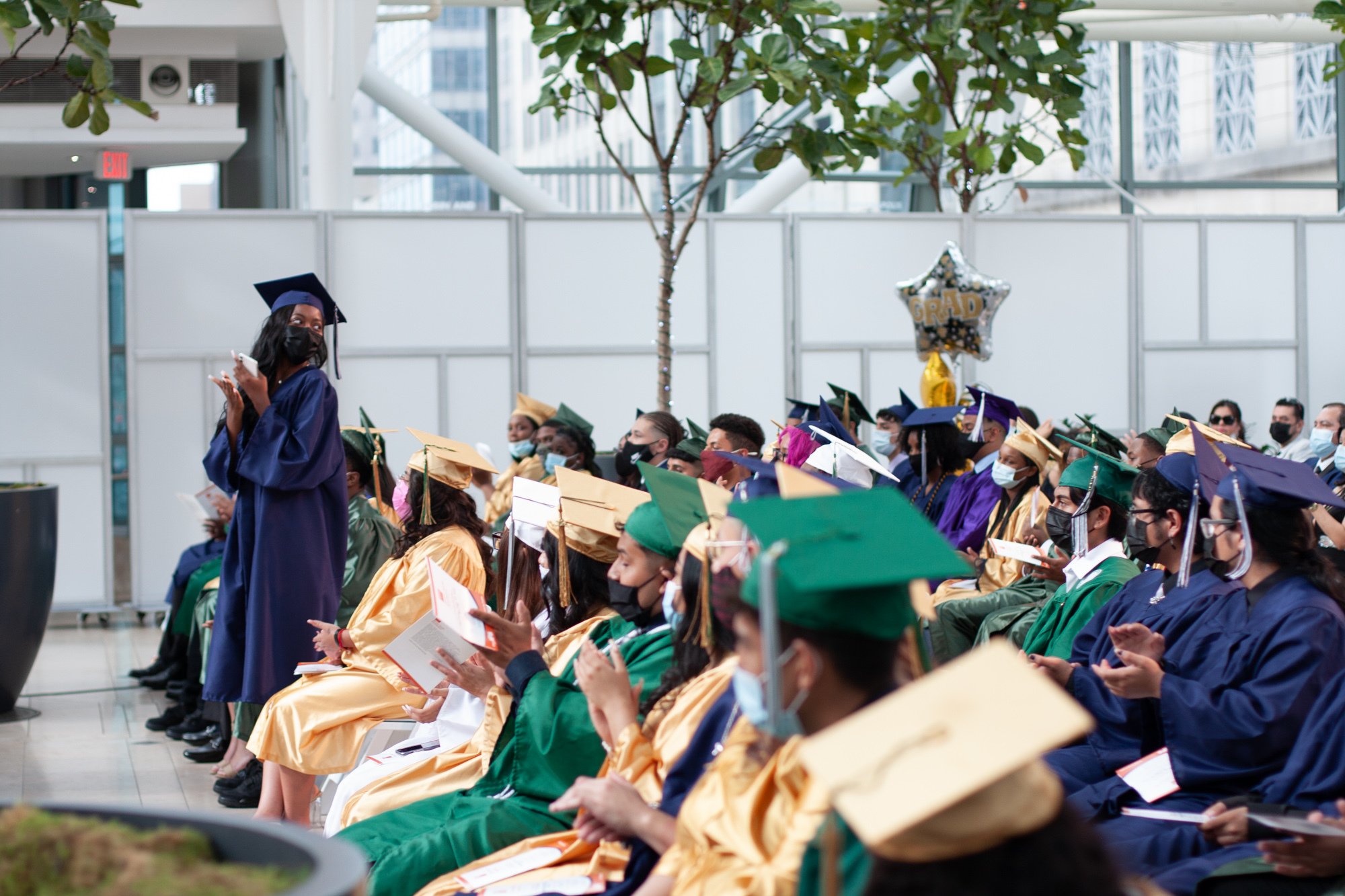 A girl in a blue cap and gown stands in a row of students wearing blue, gold and green graduation gowns and claps.