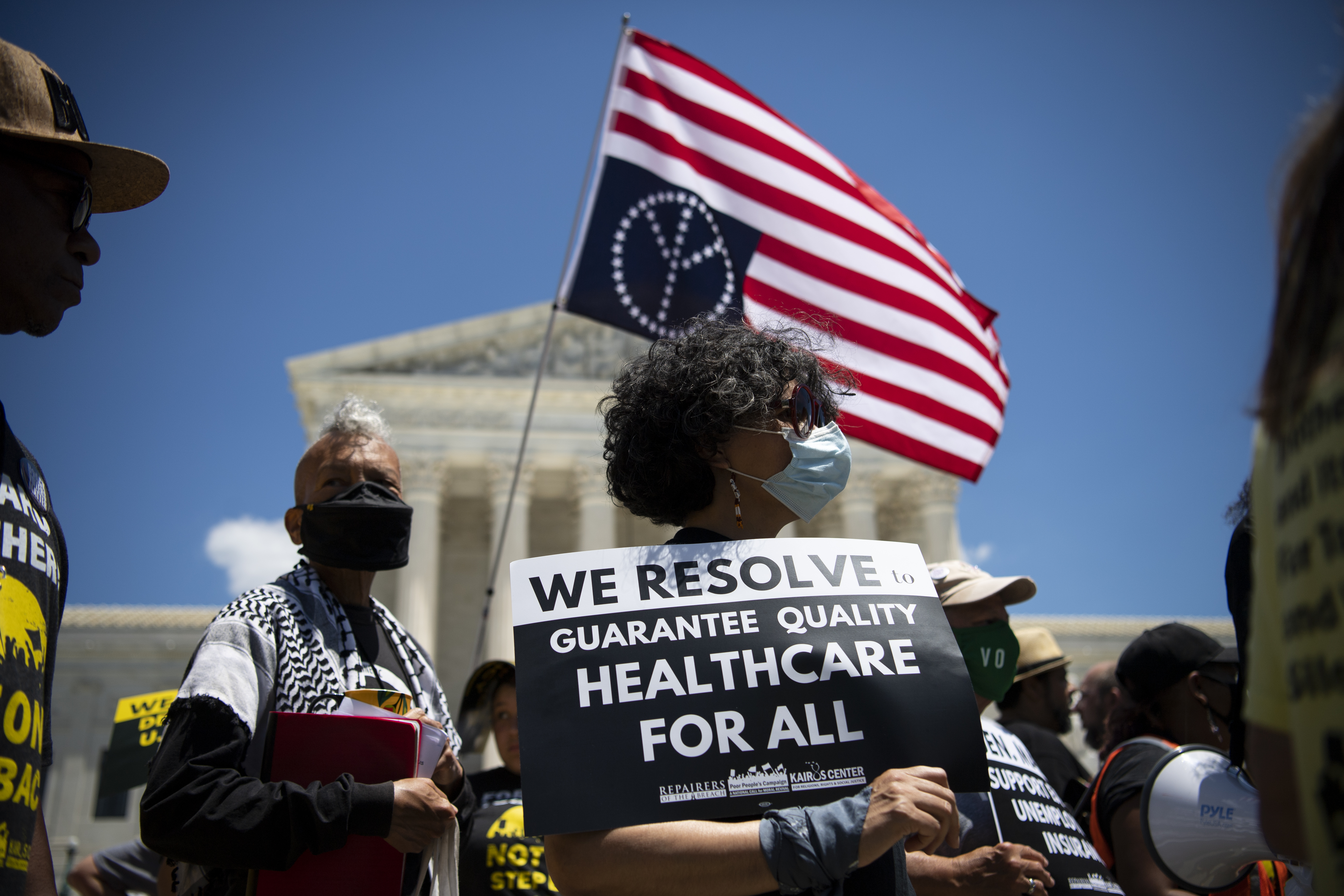 Protesters outside the Supreme Court building carry an American flag with a peace symbol on it and a sign that reads, “We resolve to guarantee quality healthcare for all.”