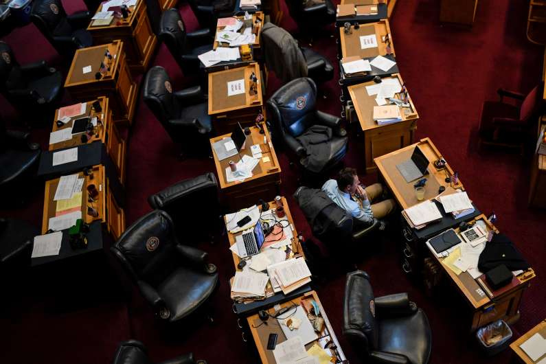 Sen. Jim Smallwood sits alone amid desks, many cluttered with papers, lined up in a semicircle in legislative chambers at the Colorado State Capitol