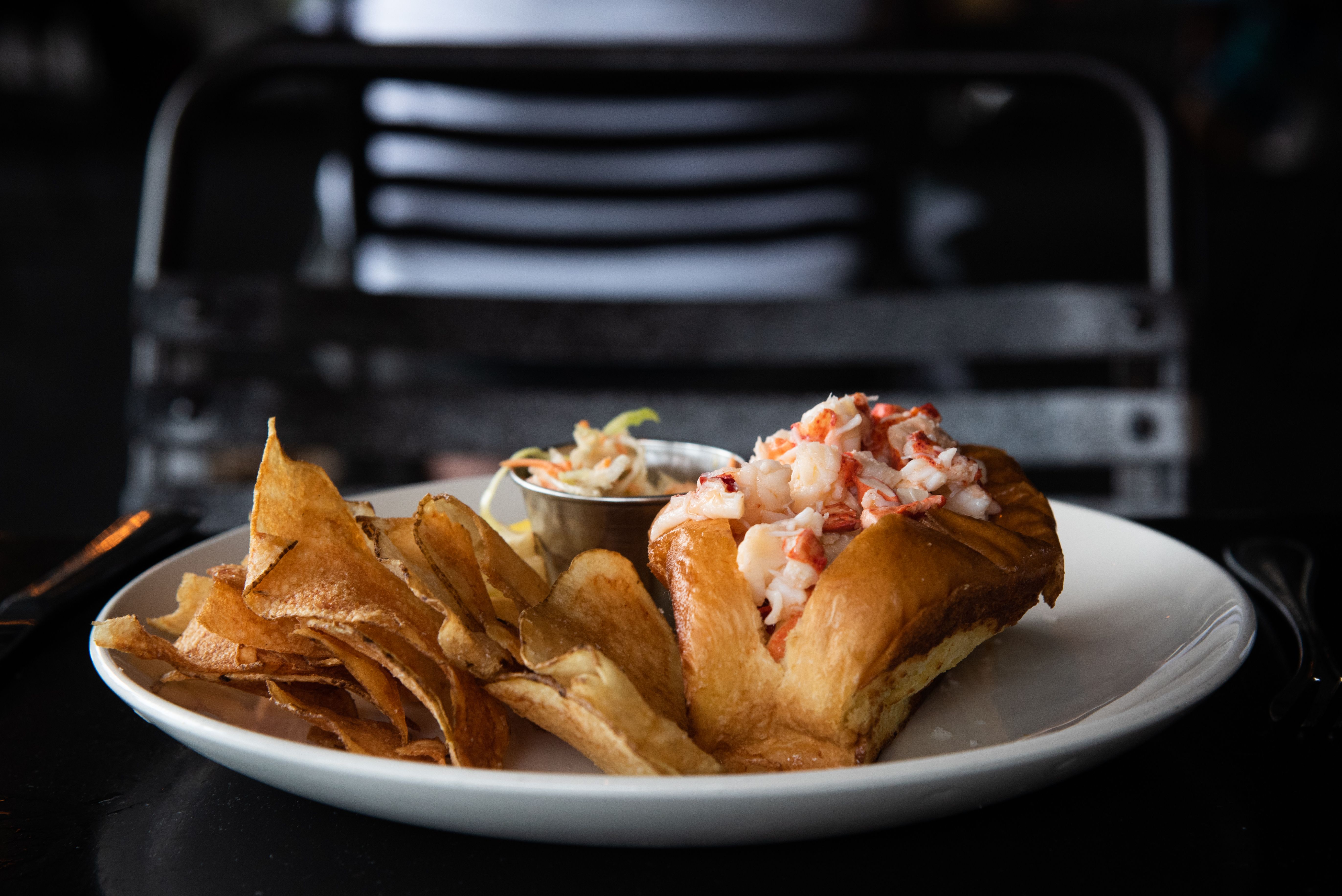 A lobster roll with a side of chips sits on a white plate on a black table
