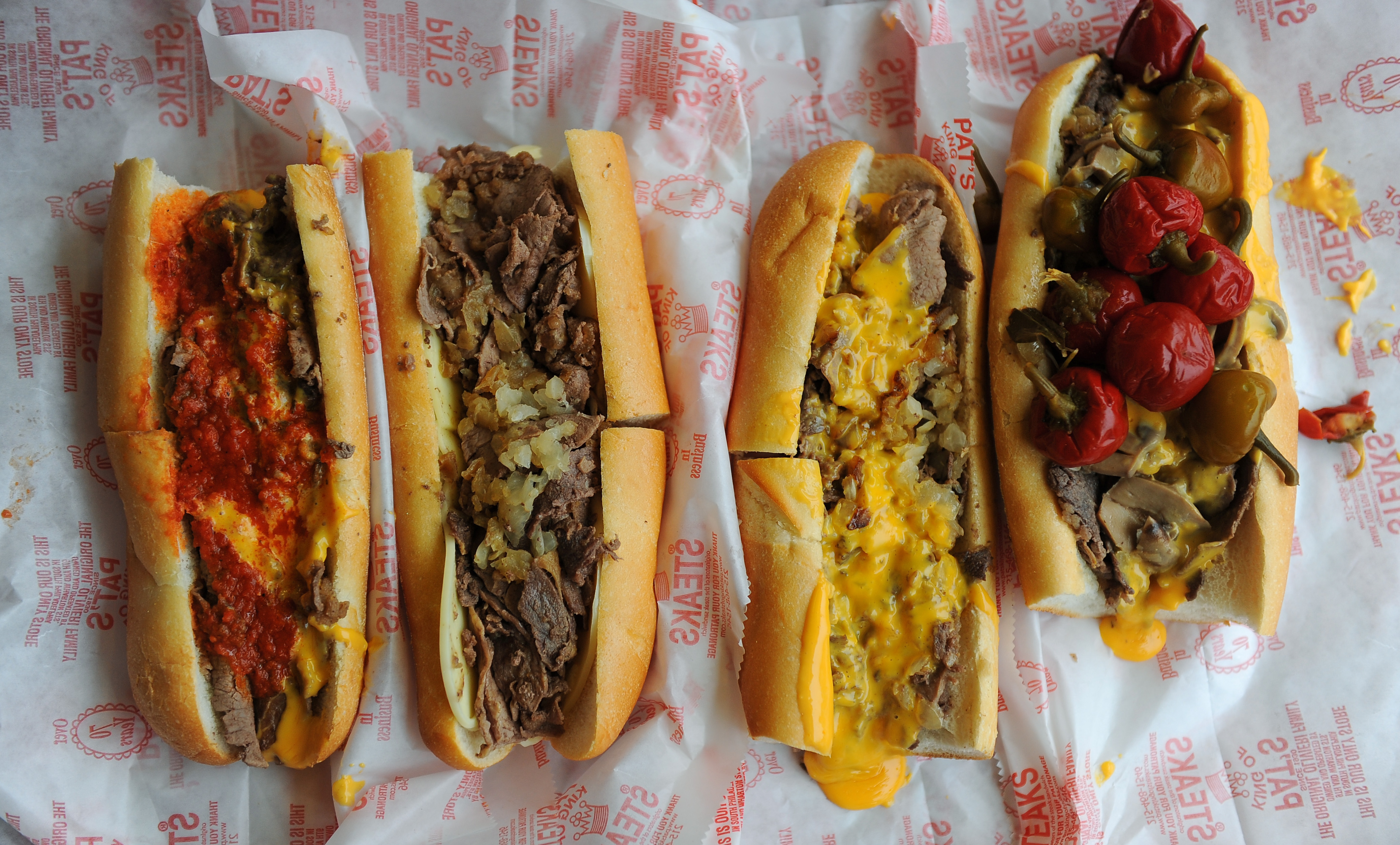 An overhead image of four cheesesteaks on white paper with red writing, one with peppers, one plain, one with Cheez Whiz, and one with marinara sauce.
