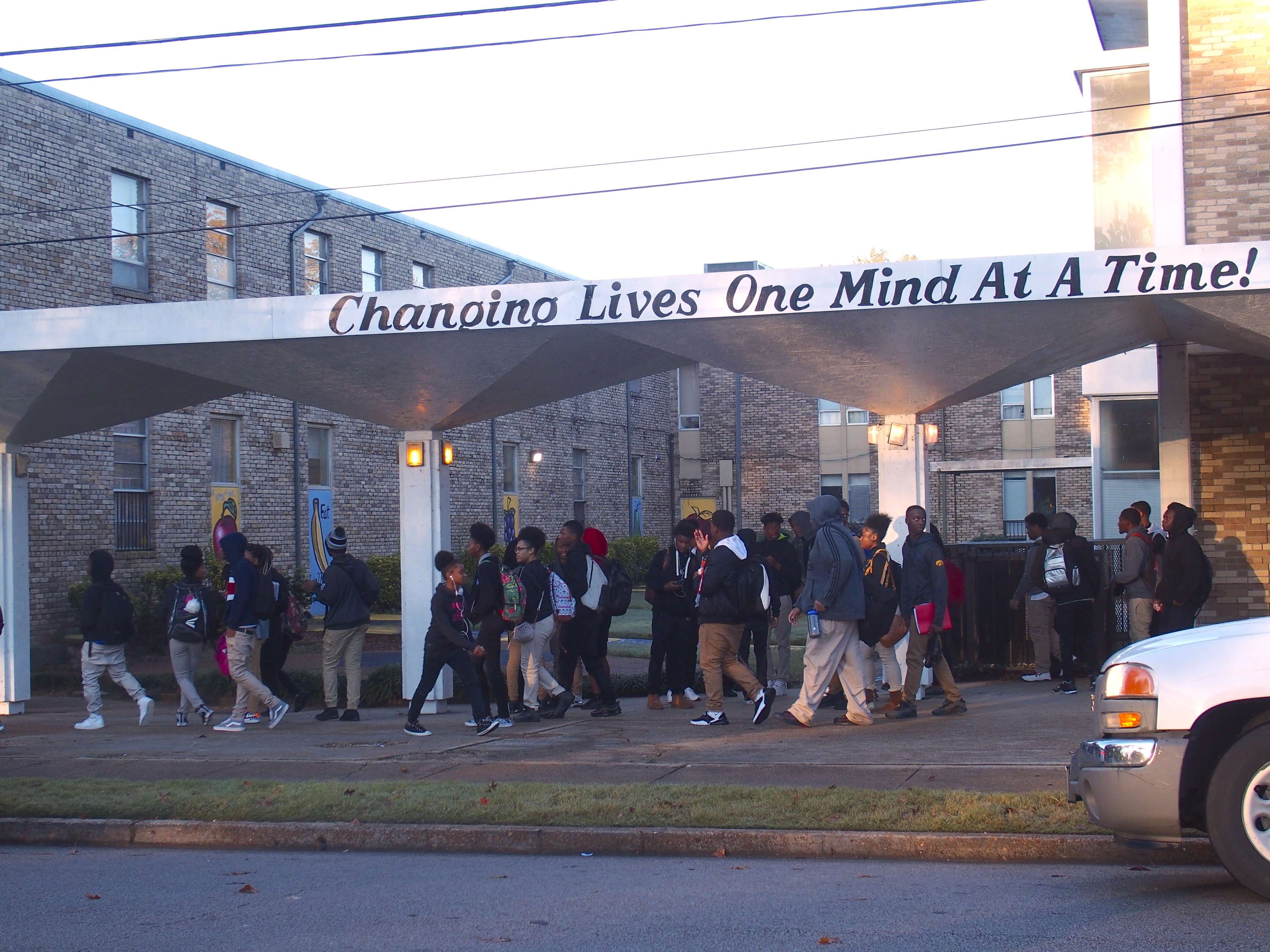 Students wearing backpacks walk under a school awning that says, “Changing lives one mind at a time!” 