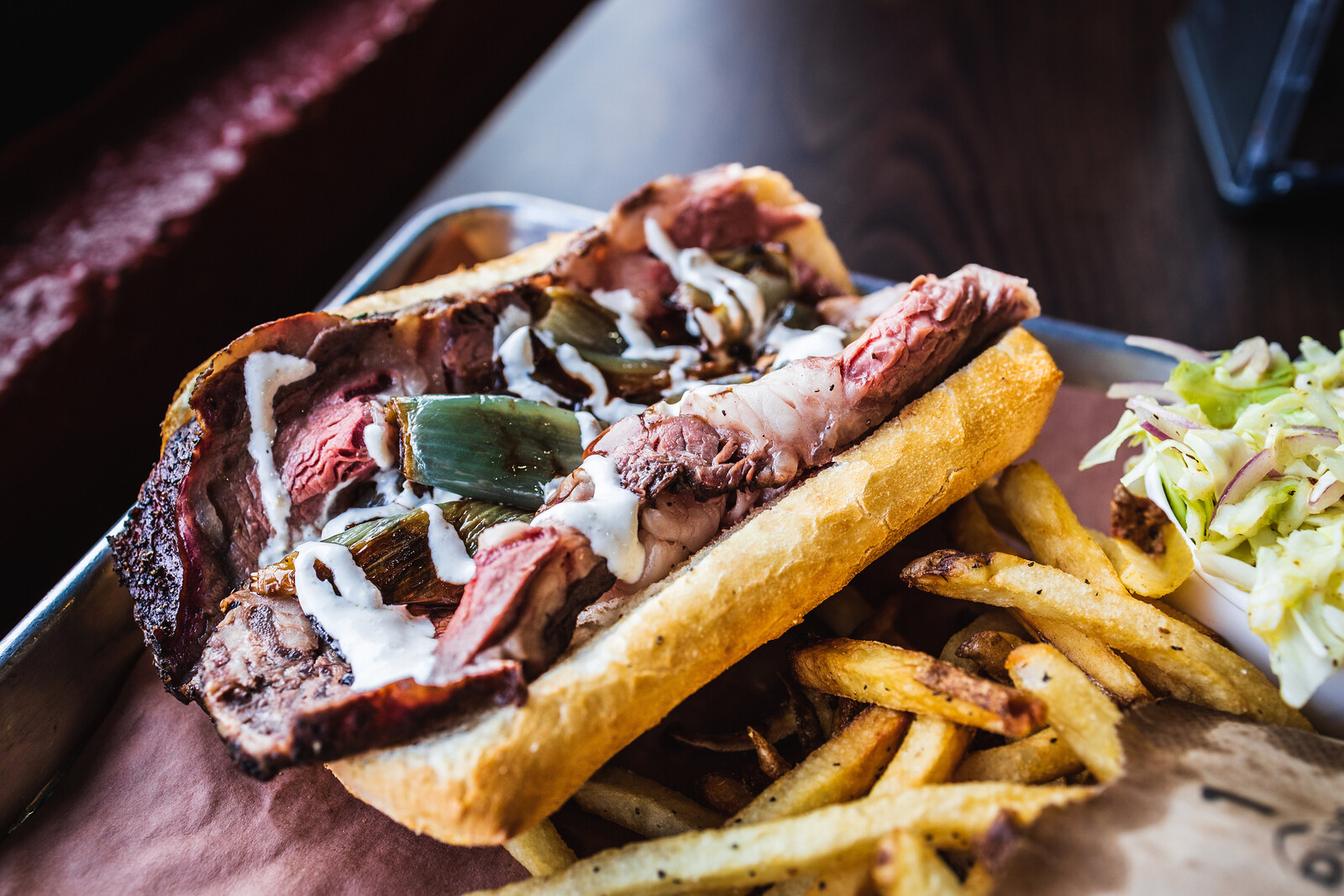 A prime rib sandwich on a French roll, topped with crispy shallot and horseradish cream, with a side of fries and coleslaw