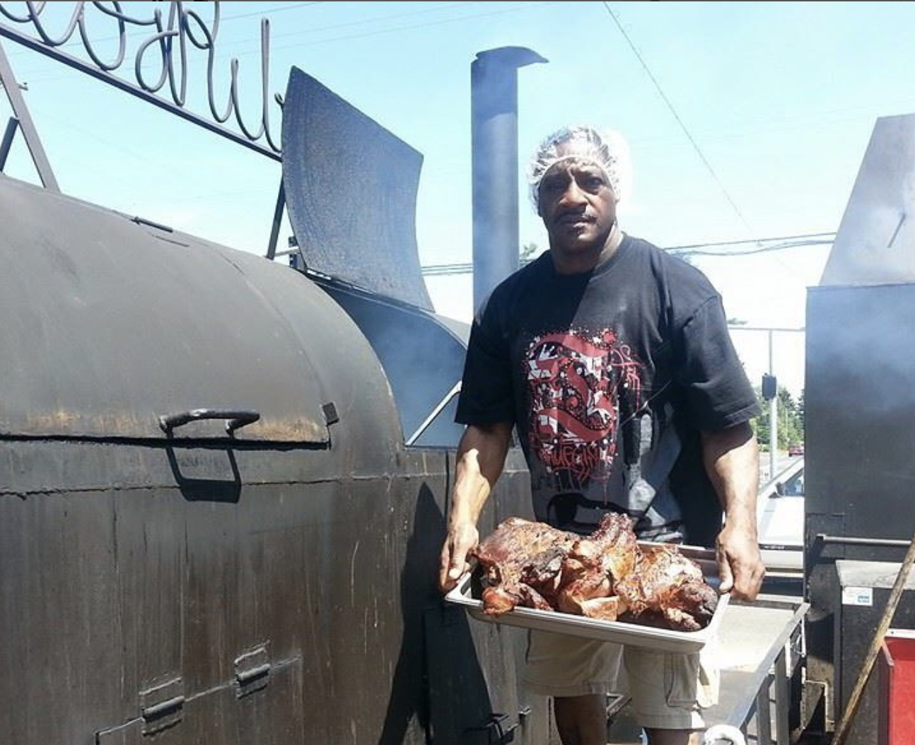 A man in a hair net holds a hunk of smoked meats next to a smoker.