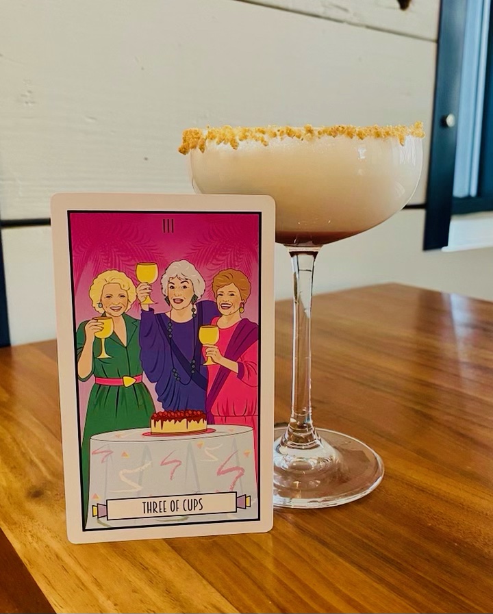 A tarot card of “The Golden Girls” next to a rum cocktail with a graham cracker crust.