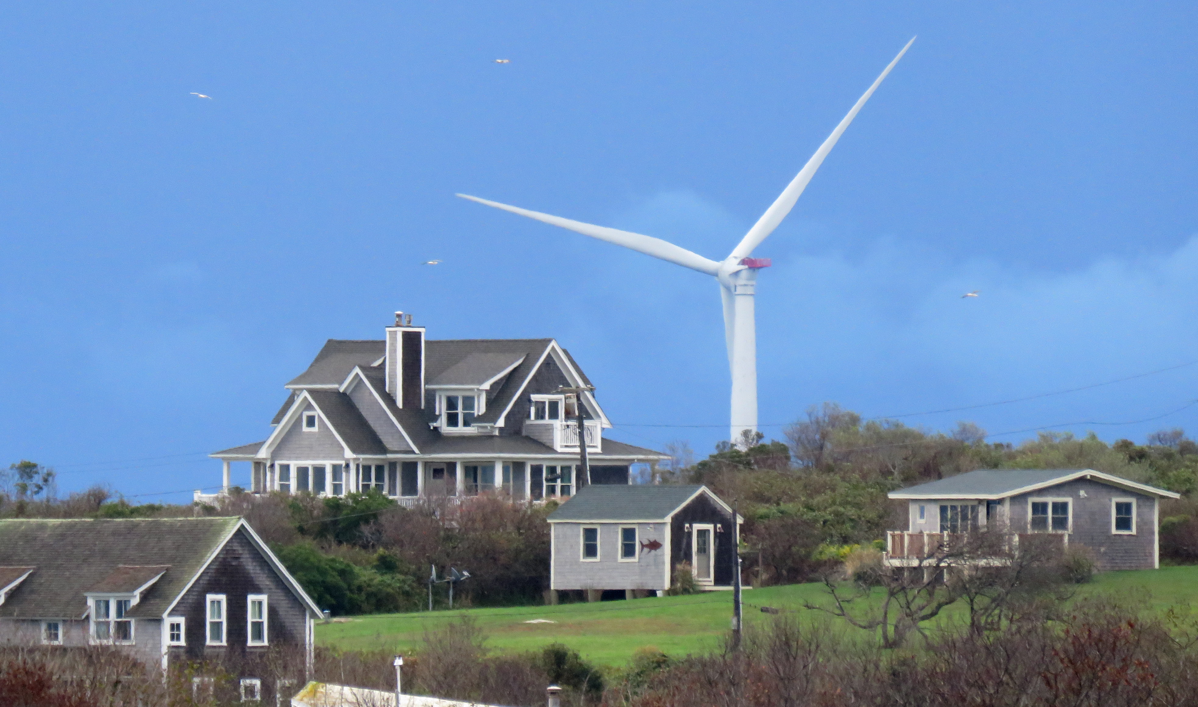The large blades of a wind turbine rise over a hill with a few houses on it. The turbine dwarfs the houses, even though it is located in the water three miles away.