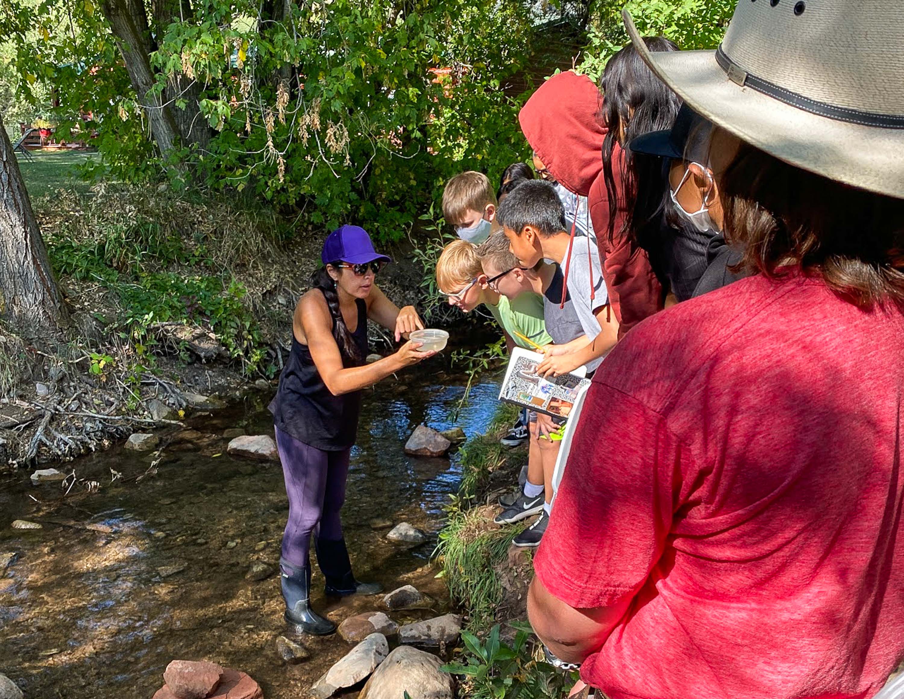 A teacher works with her students at a creek, with her children looking at something she is holding in a small container.