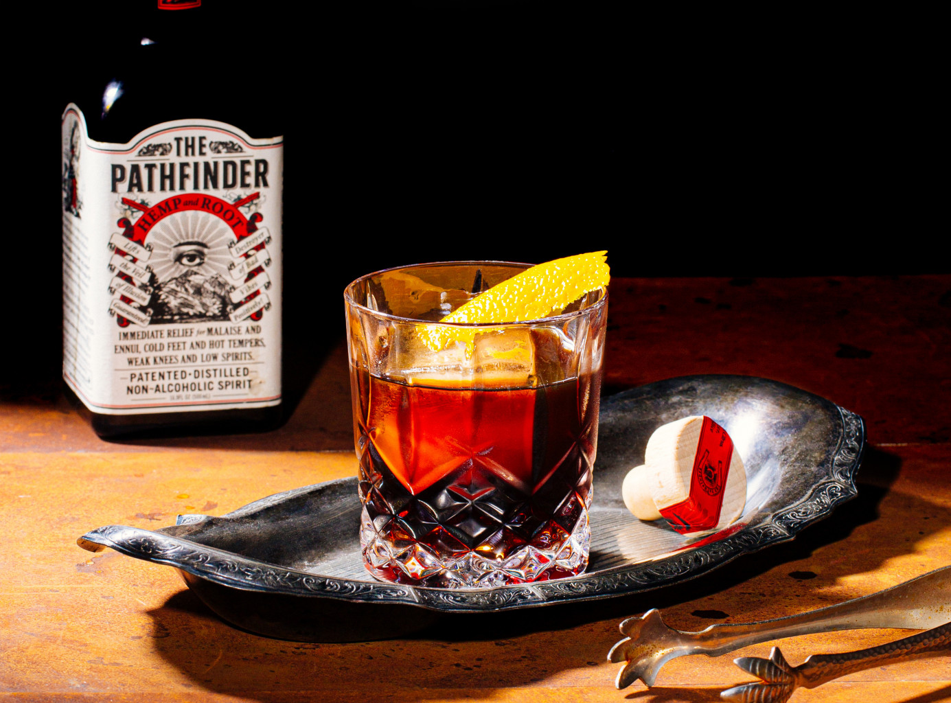 A dark, iced beverage with orange garnish sits on a silver tray. A bottle of Pathfinder is in the background.