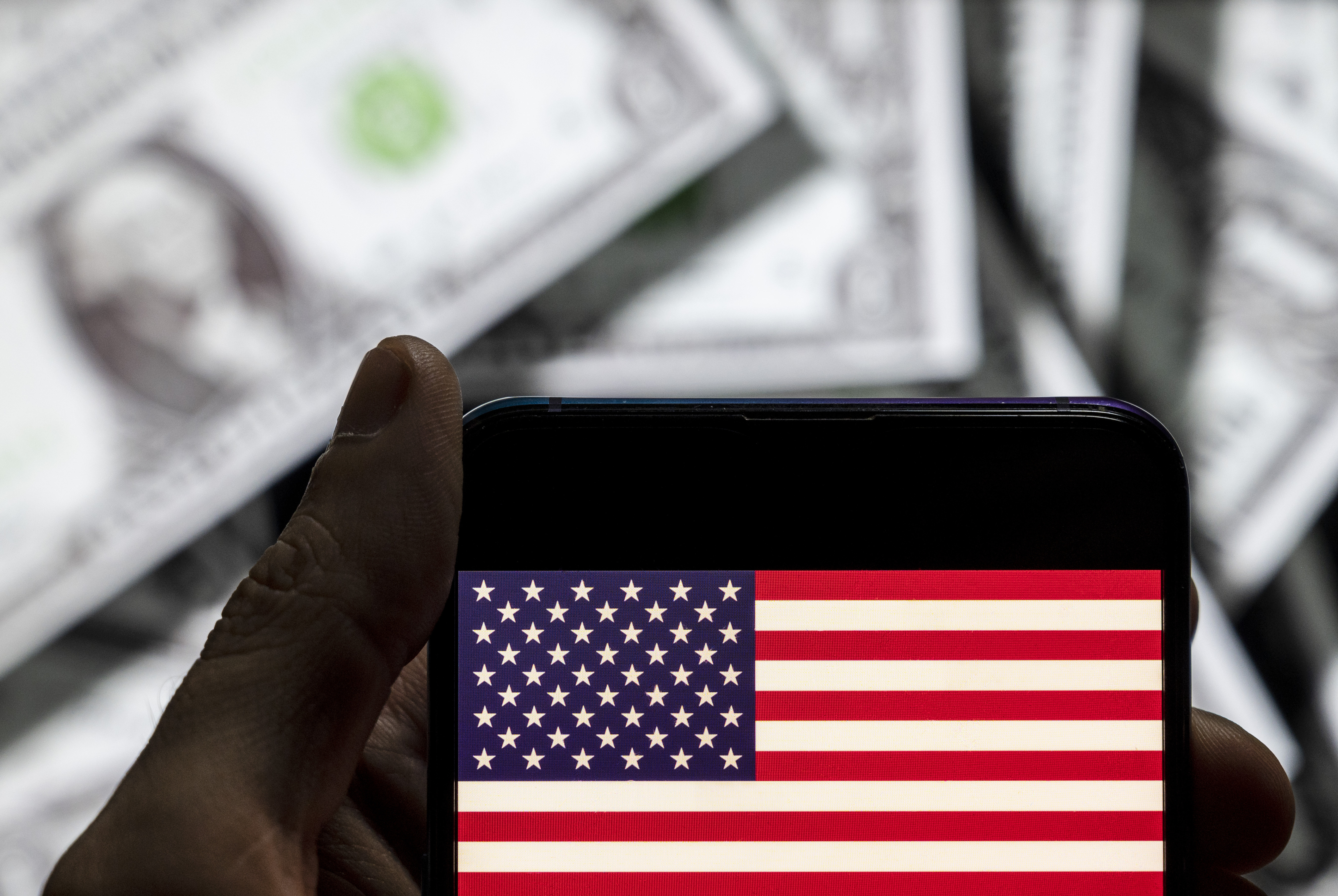 In this photo illustration the United States of America flag seen on a phone with dollar bills behind it.