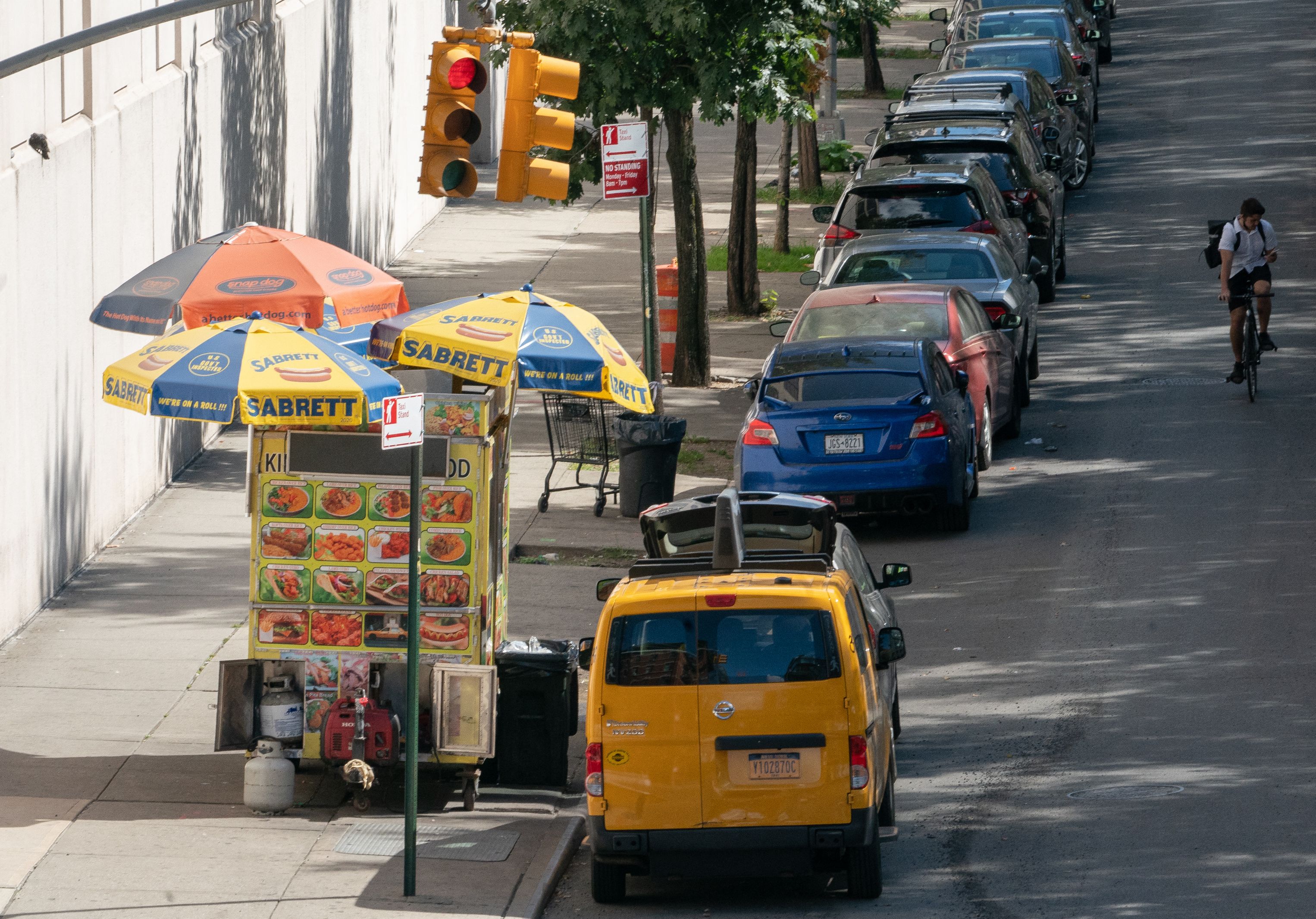 A brightly colored food cart with three umbrellas shading the cart from sun is parked on the sidewalk.