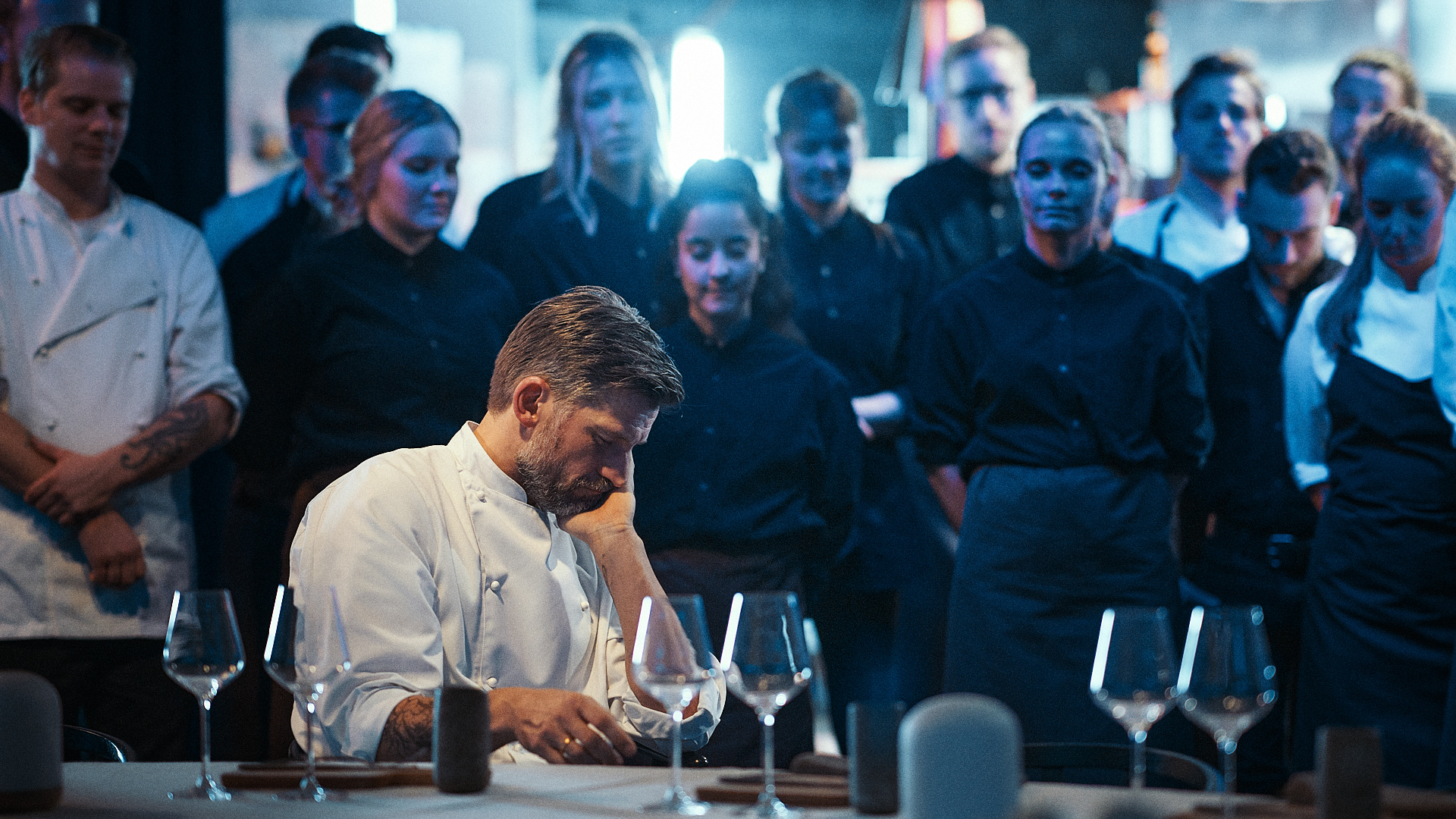 In a dark restaurant, a chef with cropped sandy brown hair and beard sits pensively at a dining table set with wine glasses, surrounded by observing waitstaff in all-black uniforms.