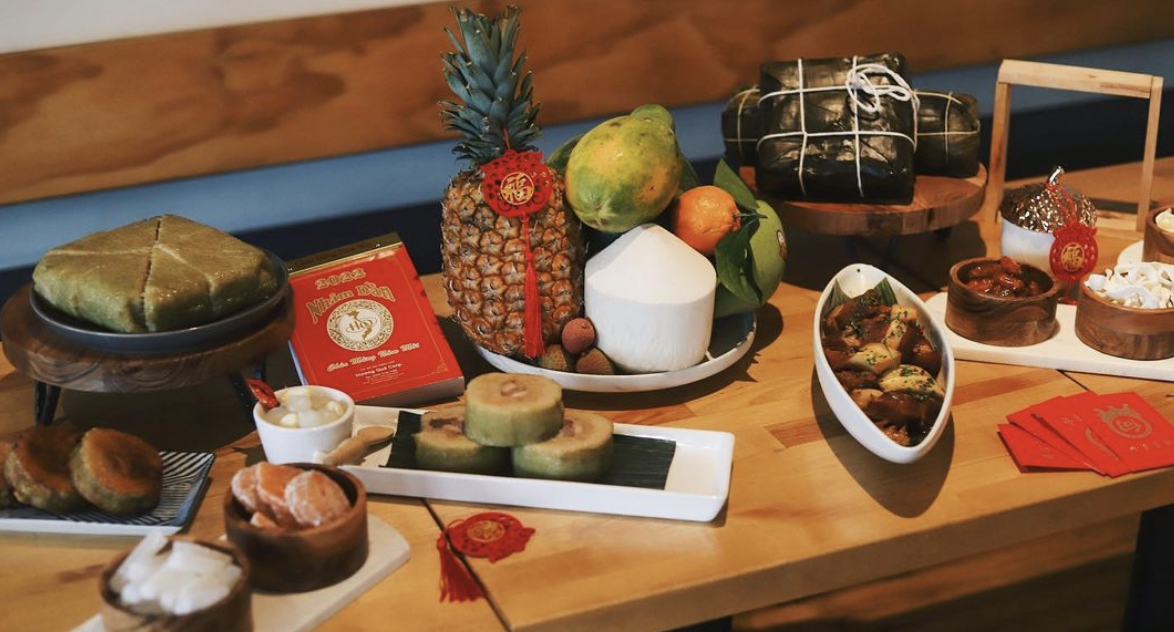 Sticky rice cakes filled with pork belly, red envelopes, and a pile of fruit on a wooden table.