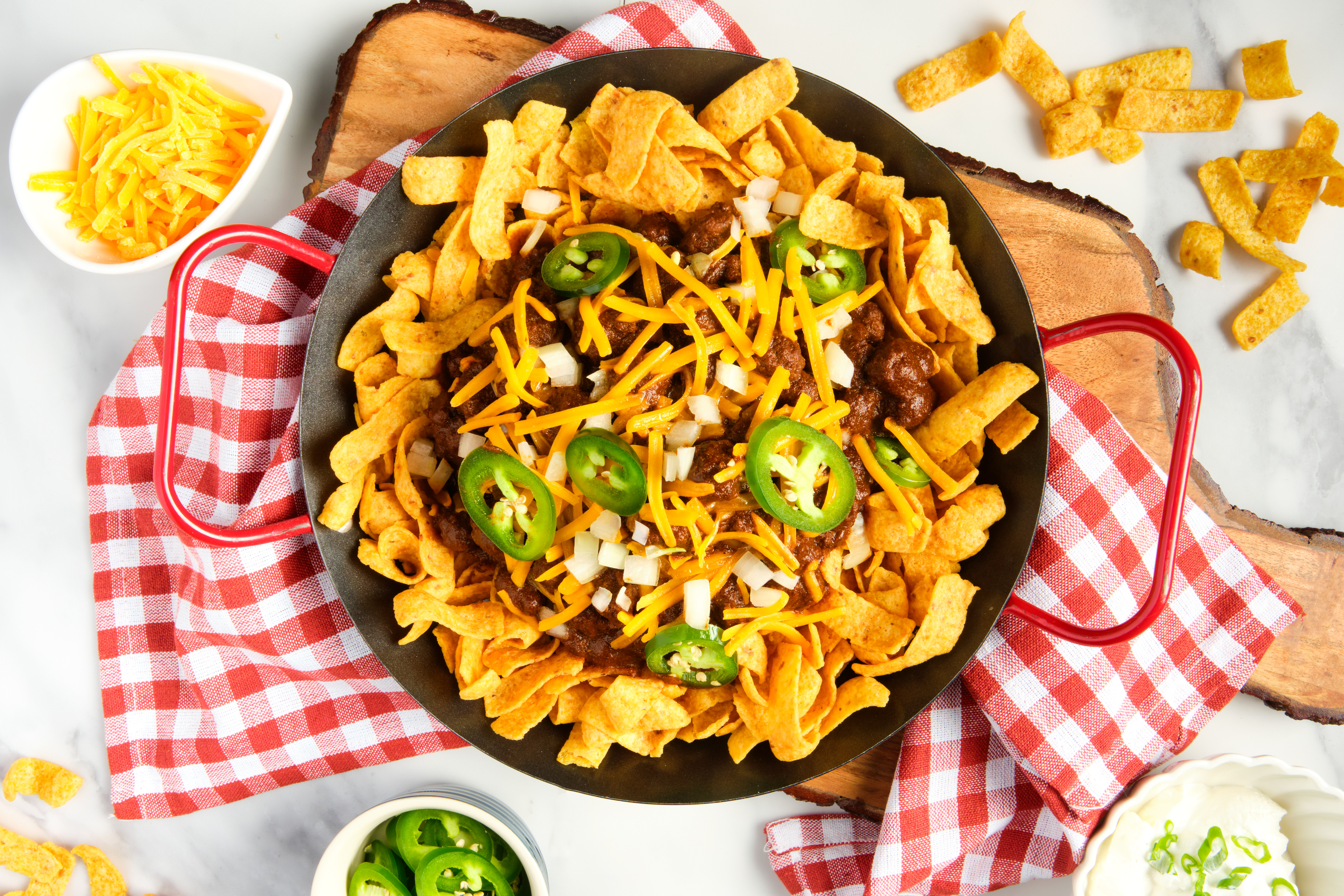 A Frito pie, topped with sliced jalapenos and shredded cheddar, is served atop a red-and-white checkered napkin and wooden cutting board. Small bowls of shredded cheese and sliced jalapenos sit nearby.