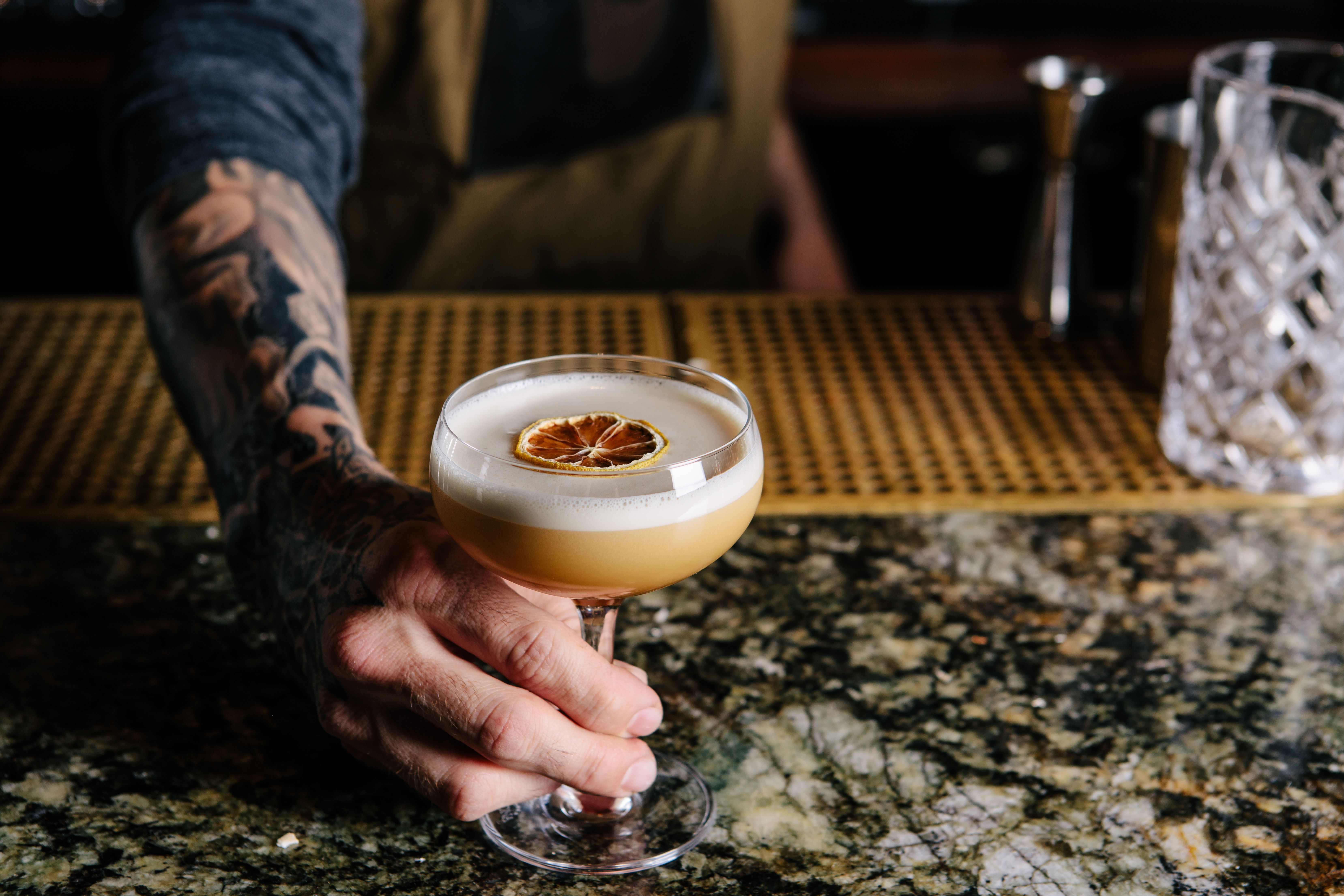 A hand pushing a coup glass filled with a frothy yellow cocktail with dried orange on top, across a bar counter. 