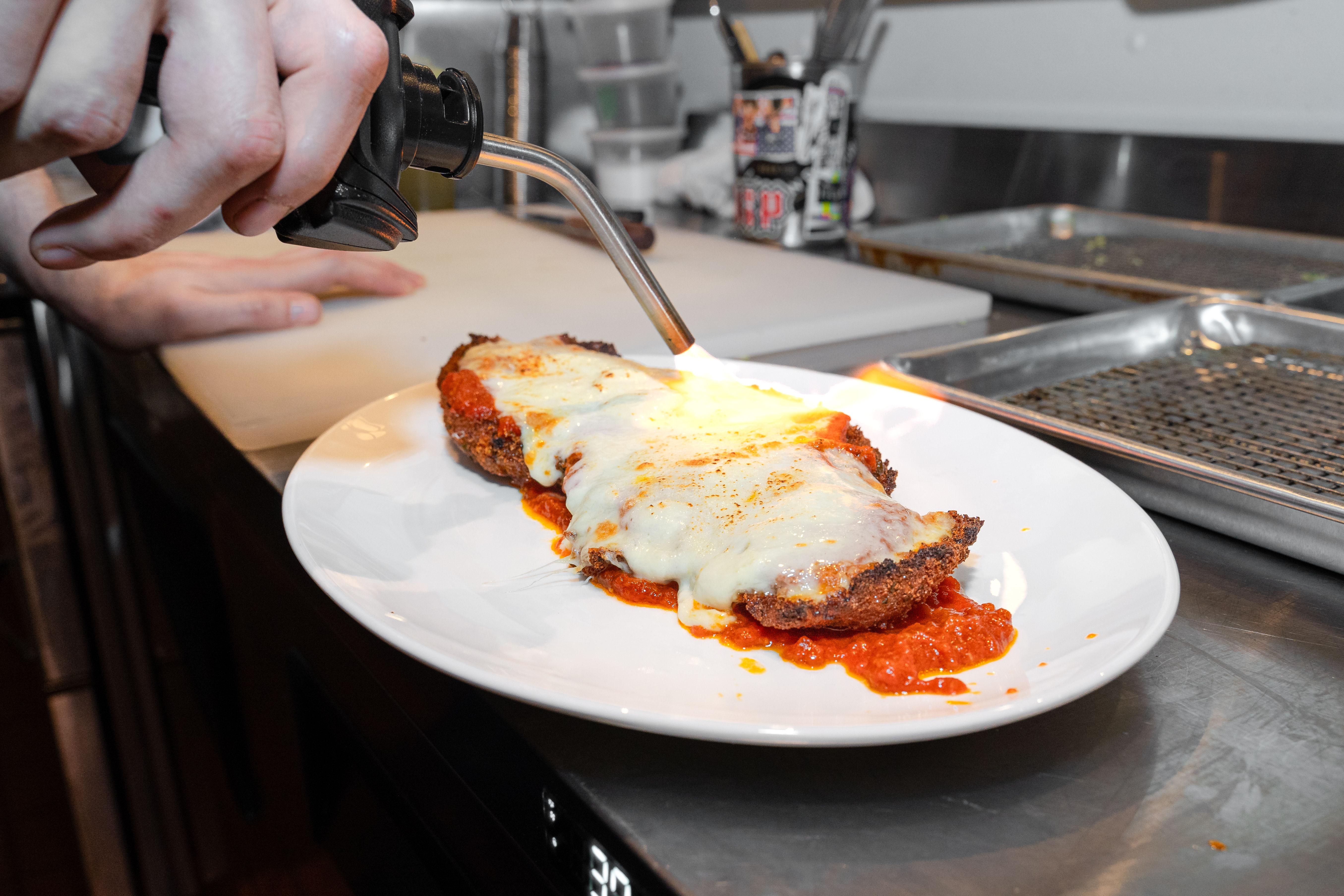 A hand holding a small kitchen blowtorch browns the cheese on top of a chicken parmigiana.
