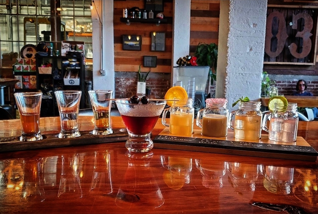 A shiny bar top holds eight distinct cocktails, each with their own style of glassware and garnish