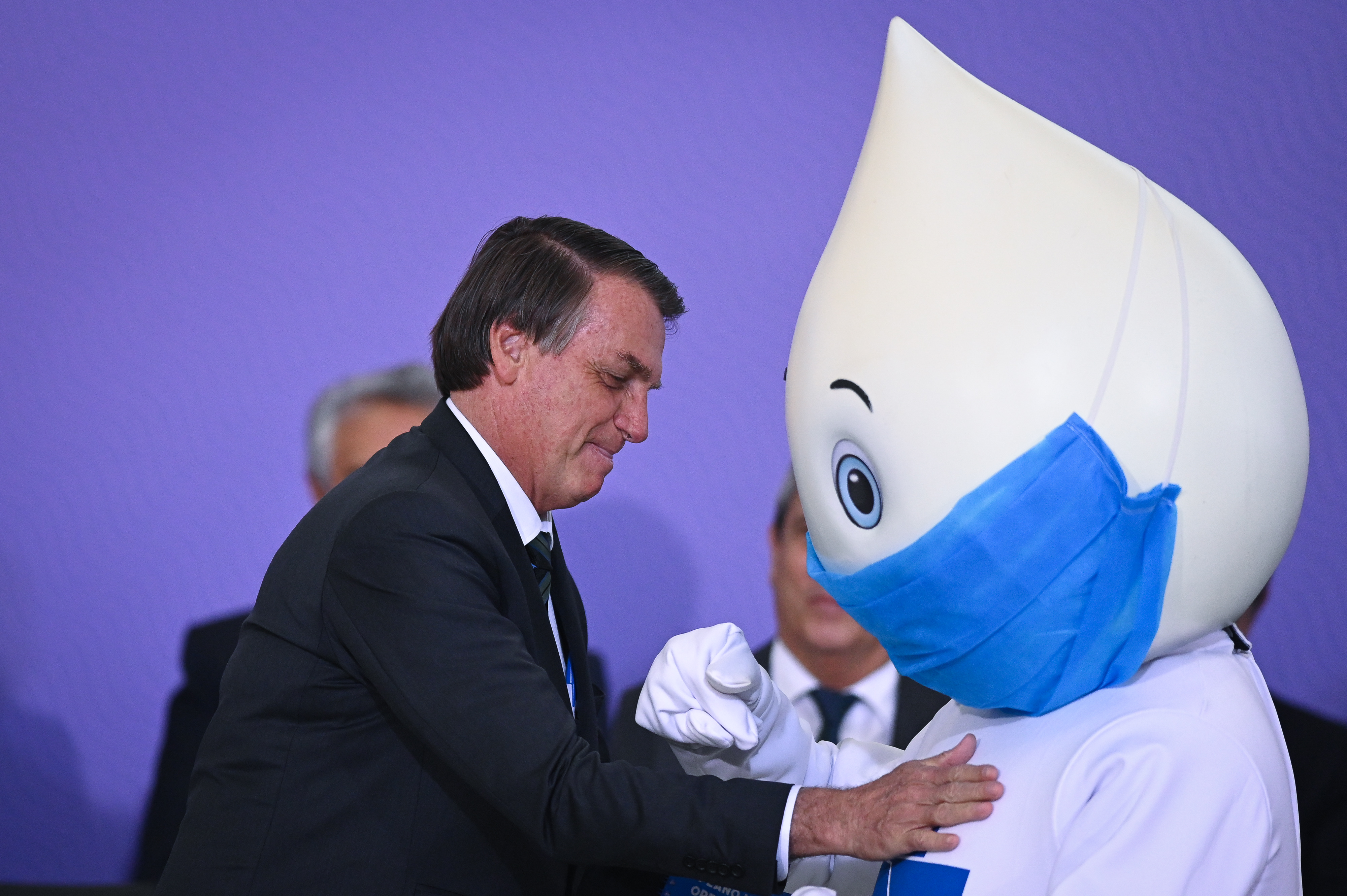 Brazilian president Jair Bolsonaro onstage with a person wearing a raindrop for a head with a huge surgical mask on its face.