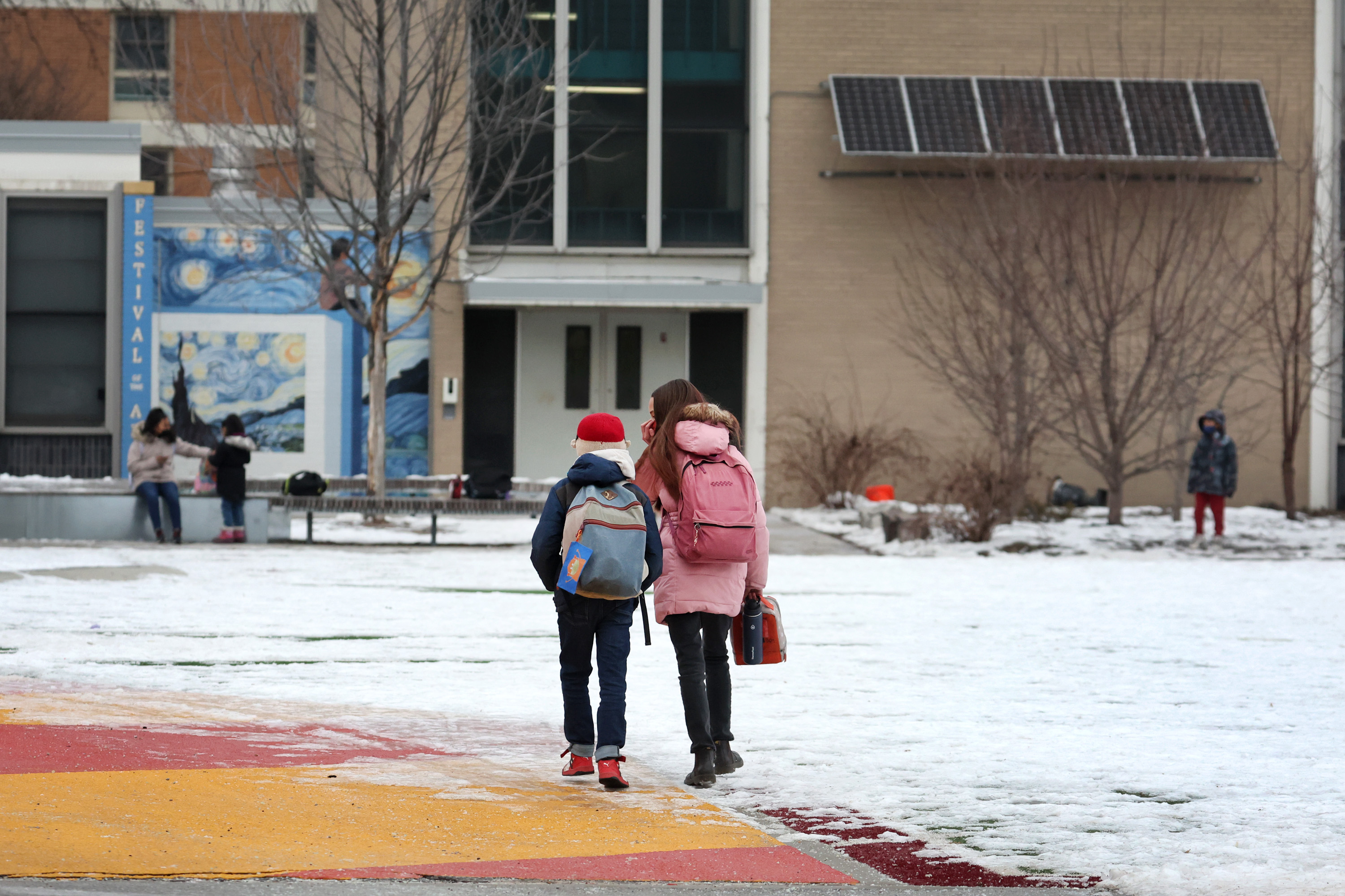 Two children, wearing backpacks, approach a school building in the snow.