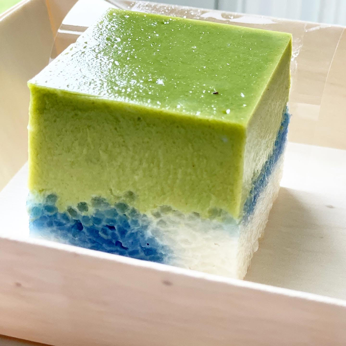 A square kuih dessert has a three layers of green, blue, and white flavors.