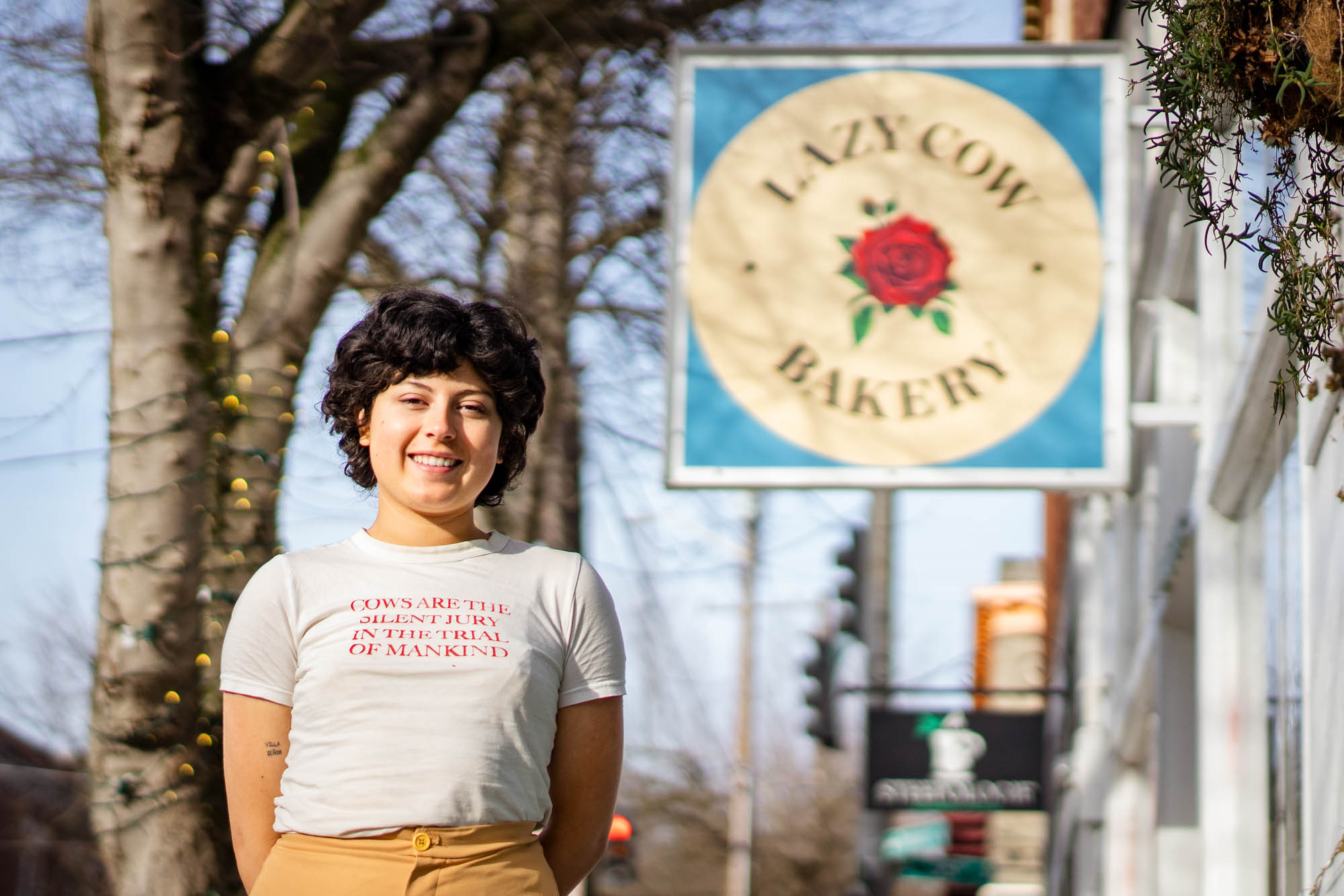 A woman stands below the sign of her bakery, Lazy Cow Bakery, on a sunny day, wearing a shirt that says “Cows are the silent jury in the trial of mankind.”