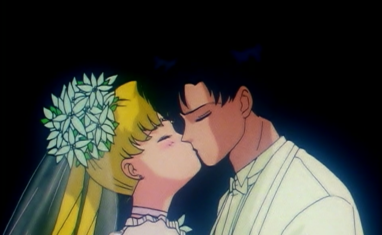 Two animated characters, Sailor Moon and Tuxedo Mask, kiss as bride and groom.
