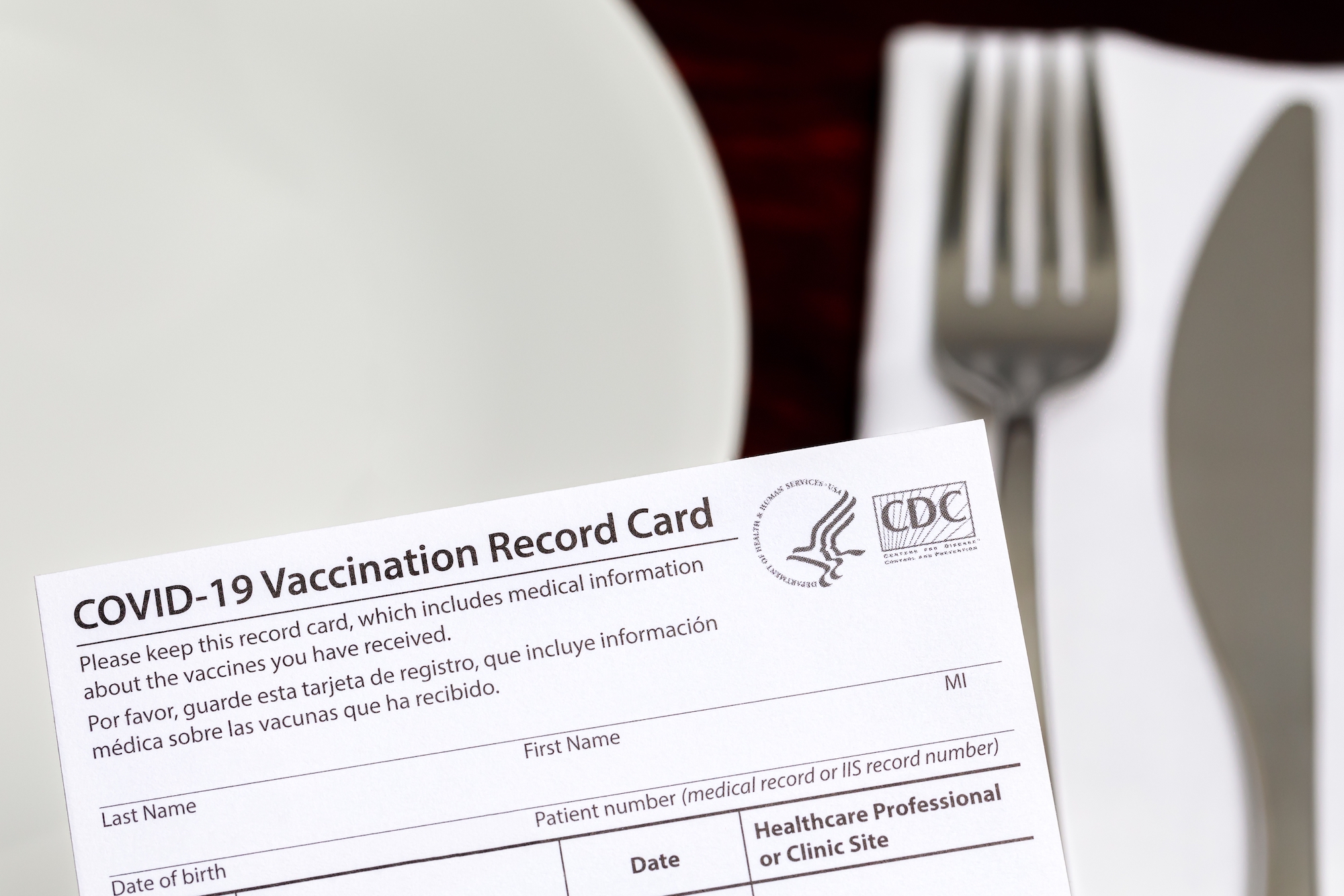 a vaccination card lies on top of a place setting of a plate, fork, knife, and napkin
