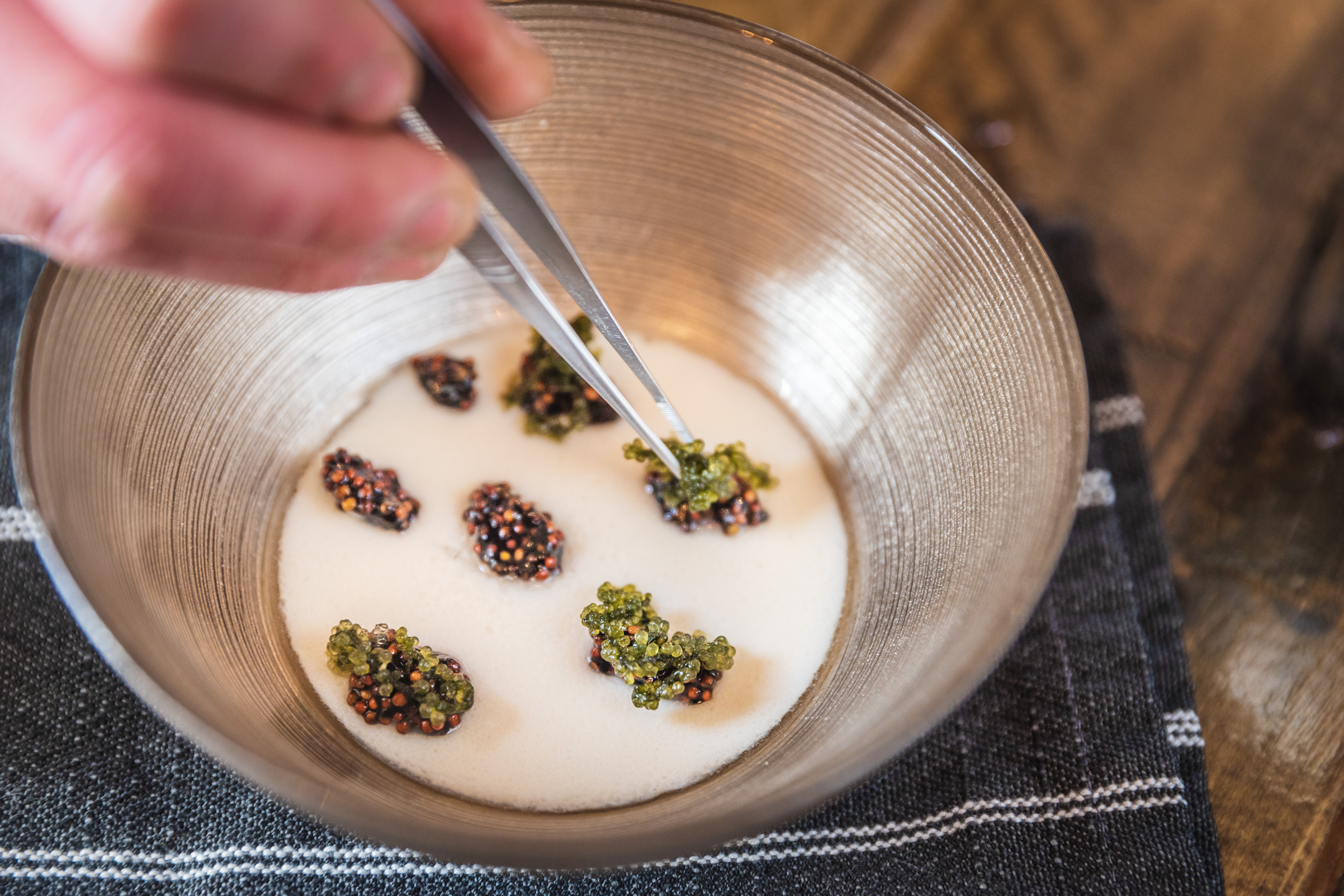 A chef’s hand uses tweezers to balance vegan caviar atop a cream-colored broth in a silver bowl.