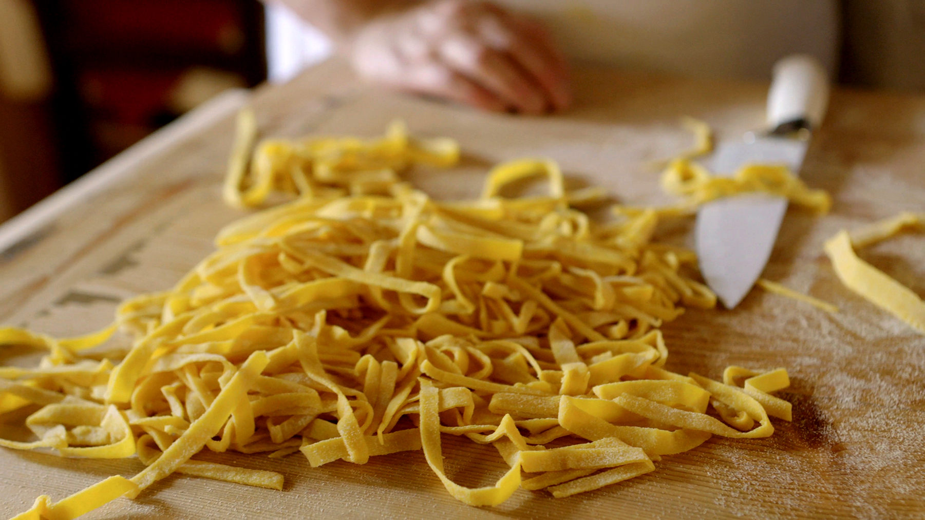 A tangle of uncooked taglietelle sits on a wooden cutting board with a chef’s knife.