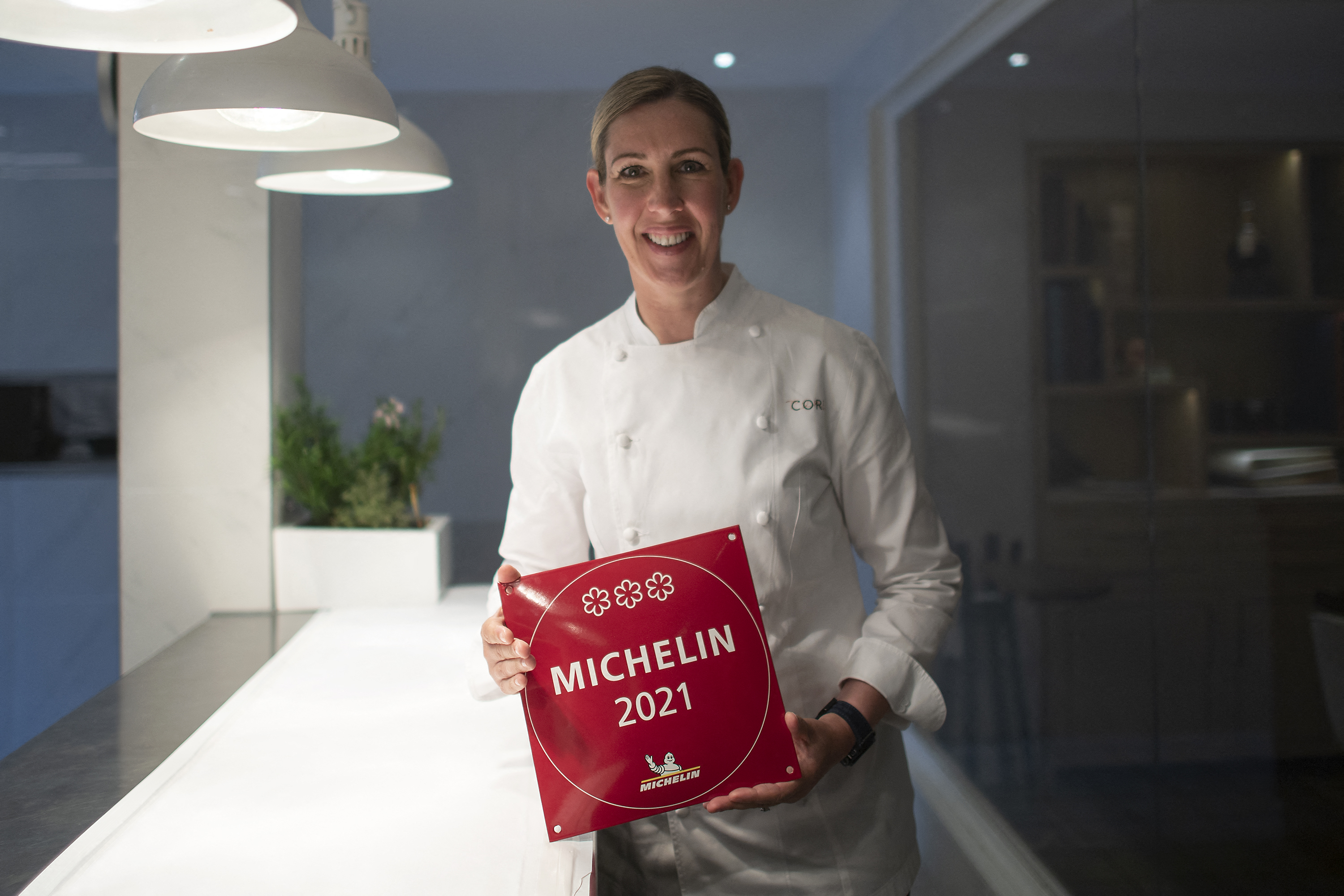Clare Smyth stands in chef’s whites, holding a red plaque that reads “Michelin 2021” in white capital letters, with three stars above the lettering
