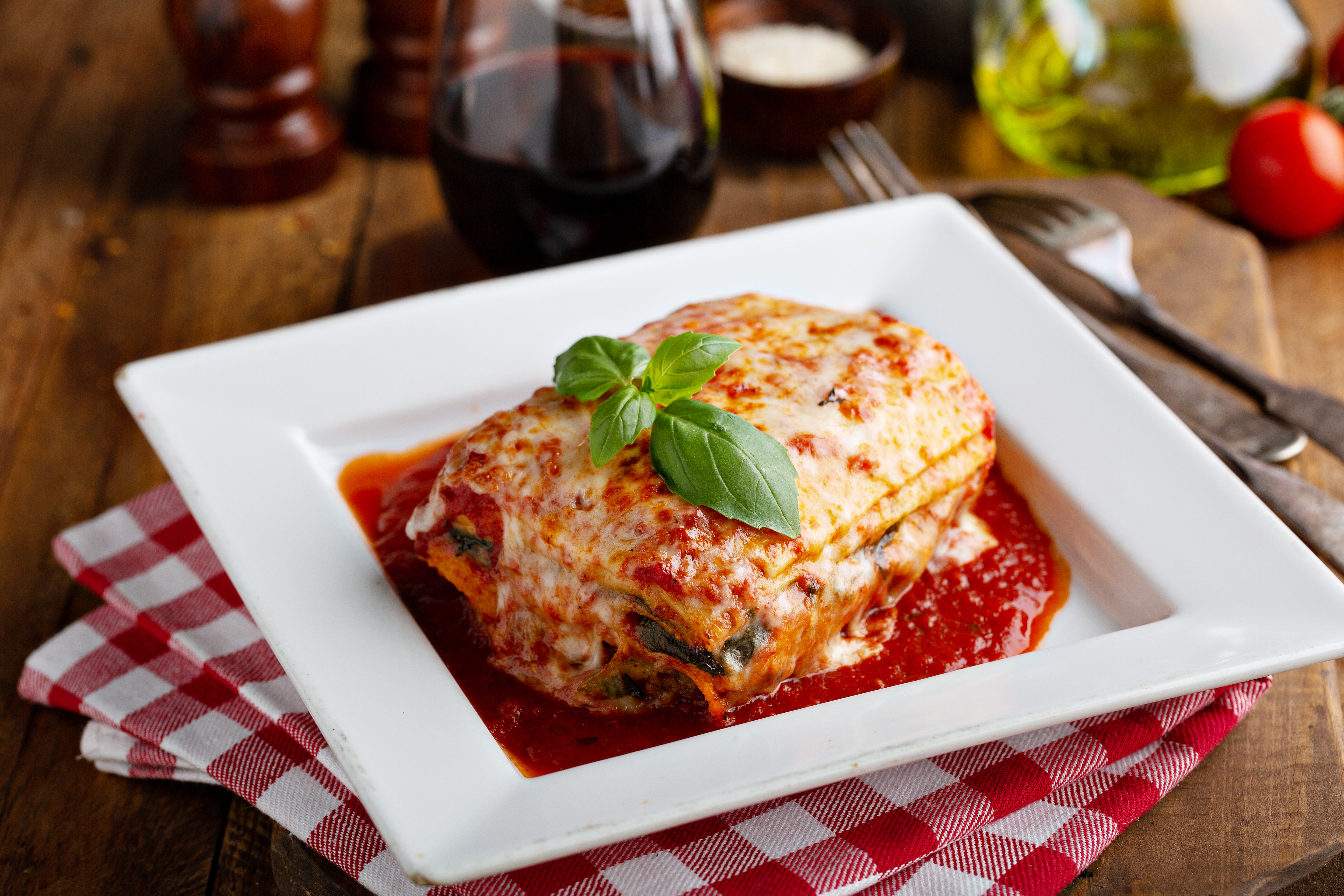 A slice of lasagna garnished with basil leaves sits in a square white dish atop a red-and-white checkered napkin.
