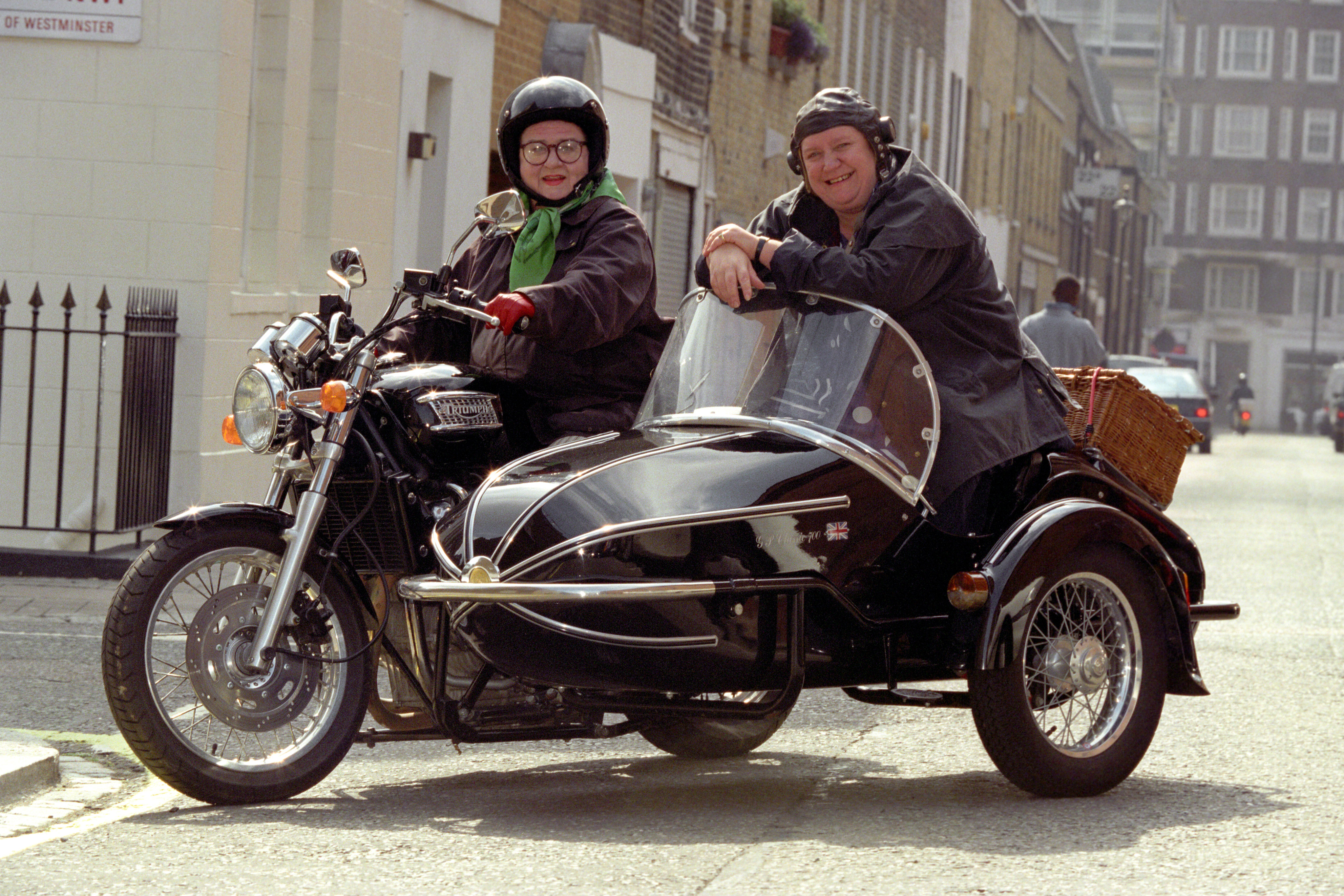Two women on a Triumph Thunderbird motorcycle, one in the driver’s seat and one in the side car, both wearing helmets, on a London residential street.