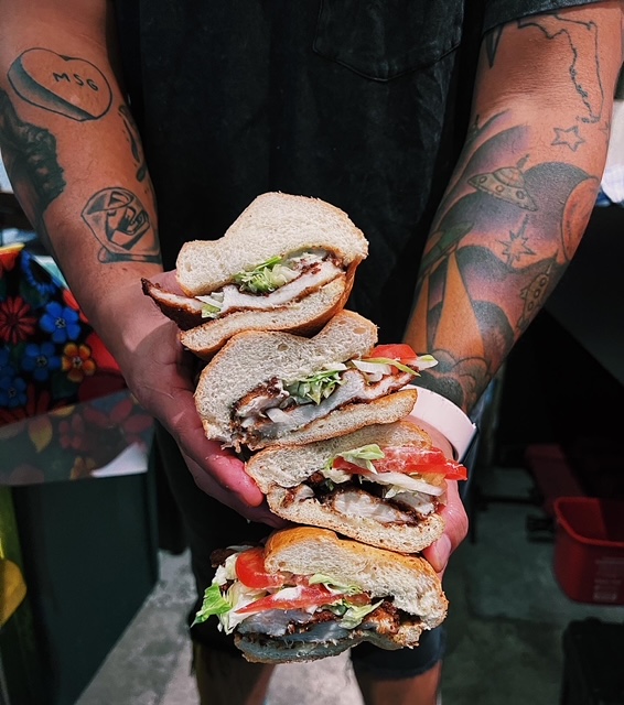 A man with tattoos holds a stack of four fried chicken cutlet sandwiches.