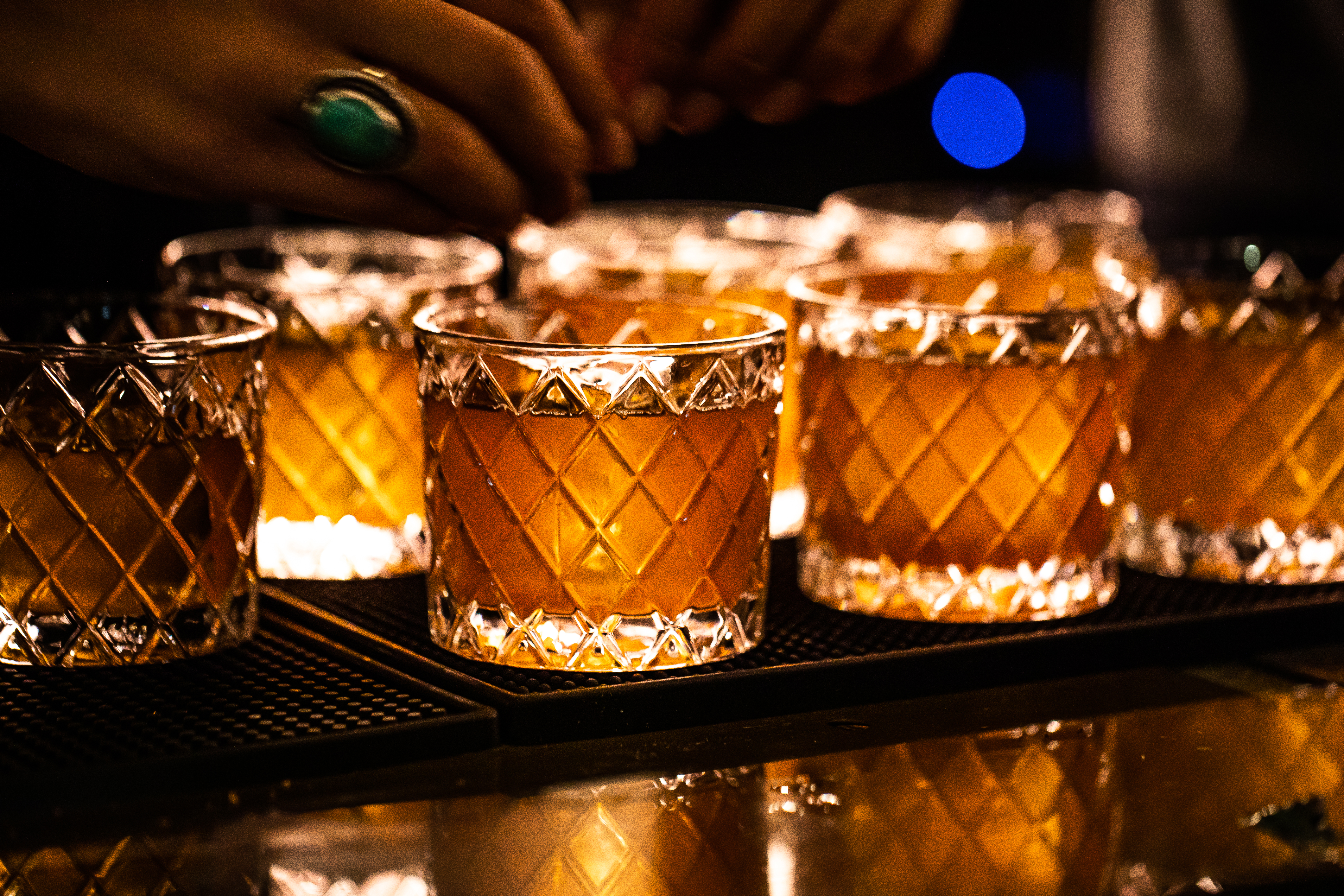 A bar top holds seven rocks glasses with diamond patterns. The drinks within are golden brown with a large ice cube.