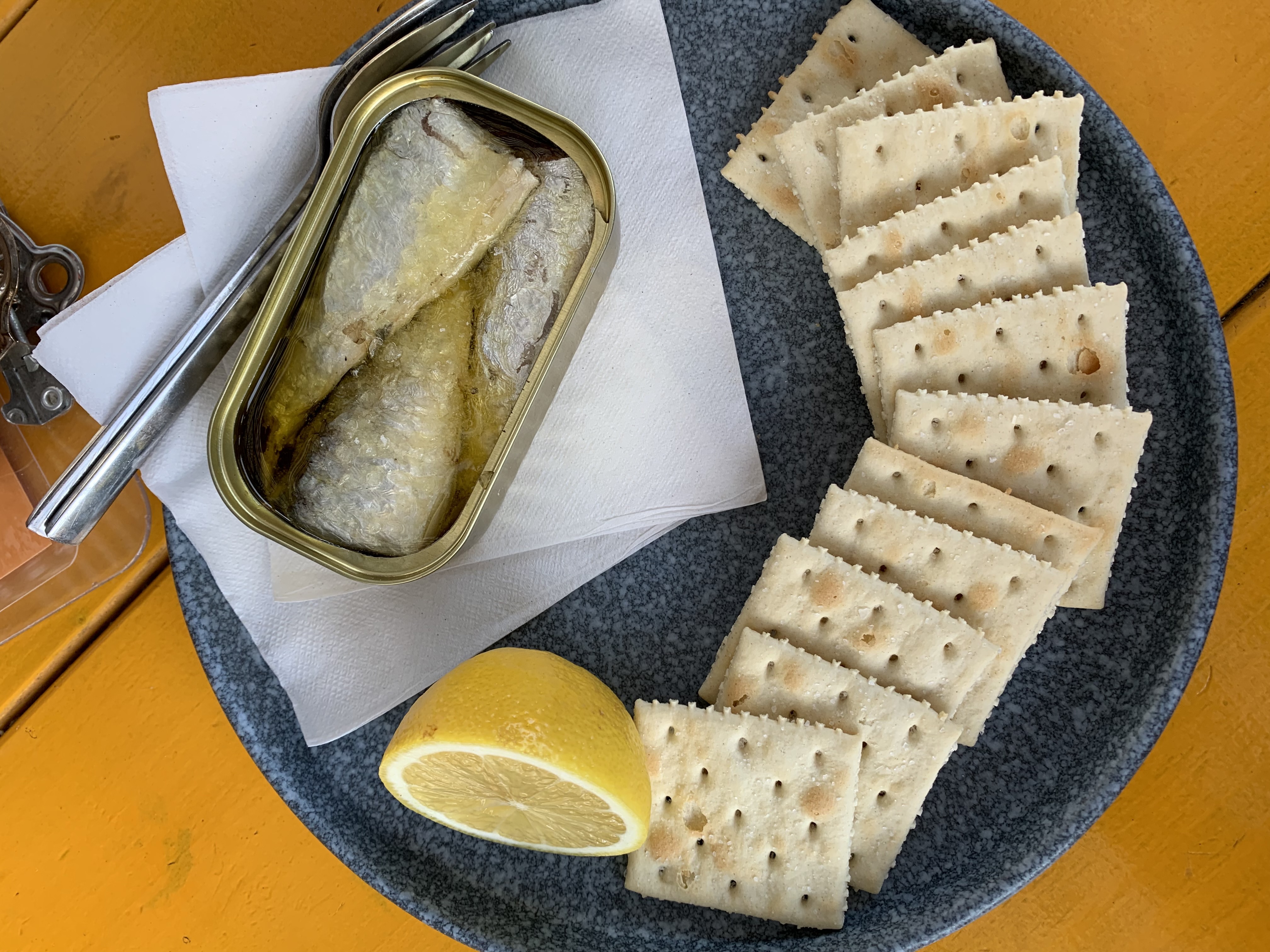 A dark-gray plate of tinned sardines, a row of crackers, and a half-lemon on an orange table.