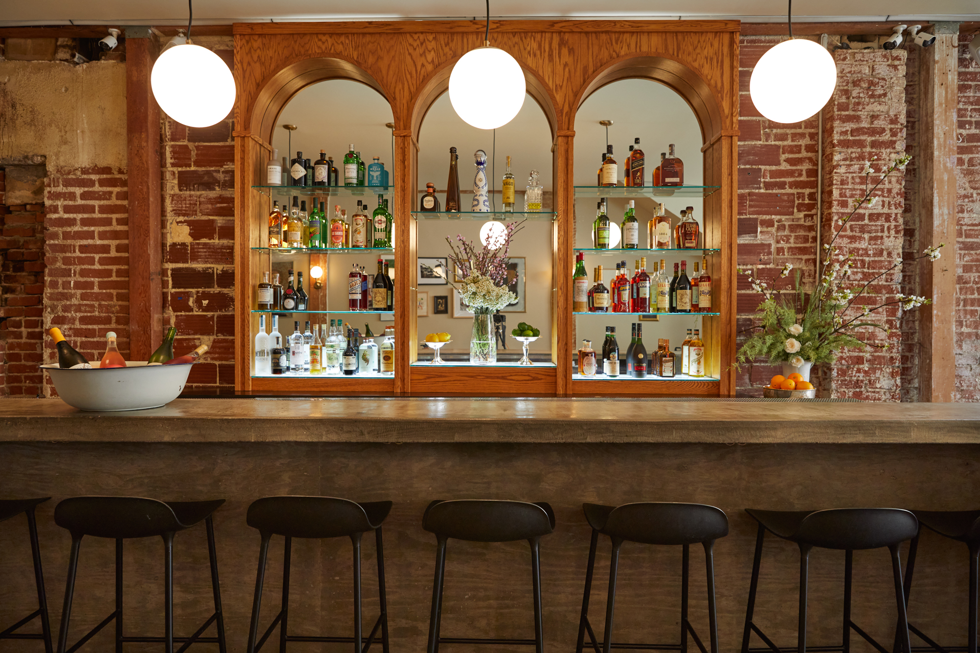 A bar with exposed brick walls, barstools, and an arched back with bottles of alcohol at Coucou restaurant in Venice, California.