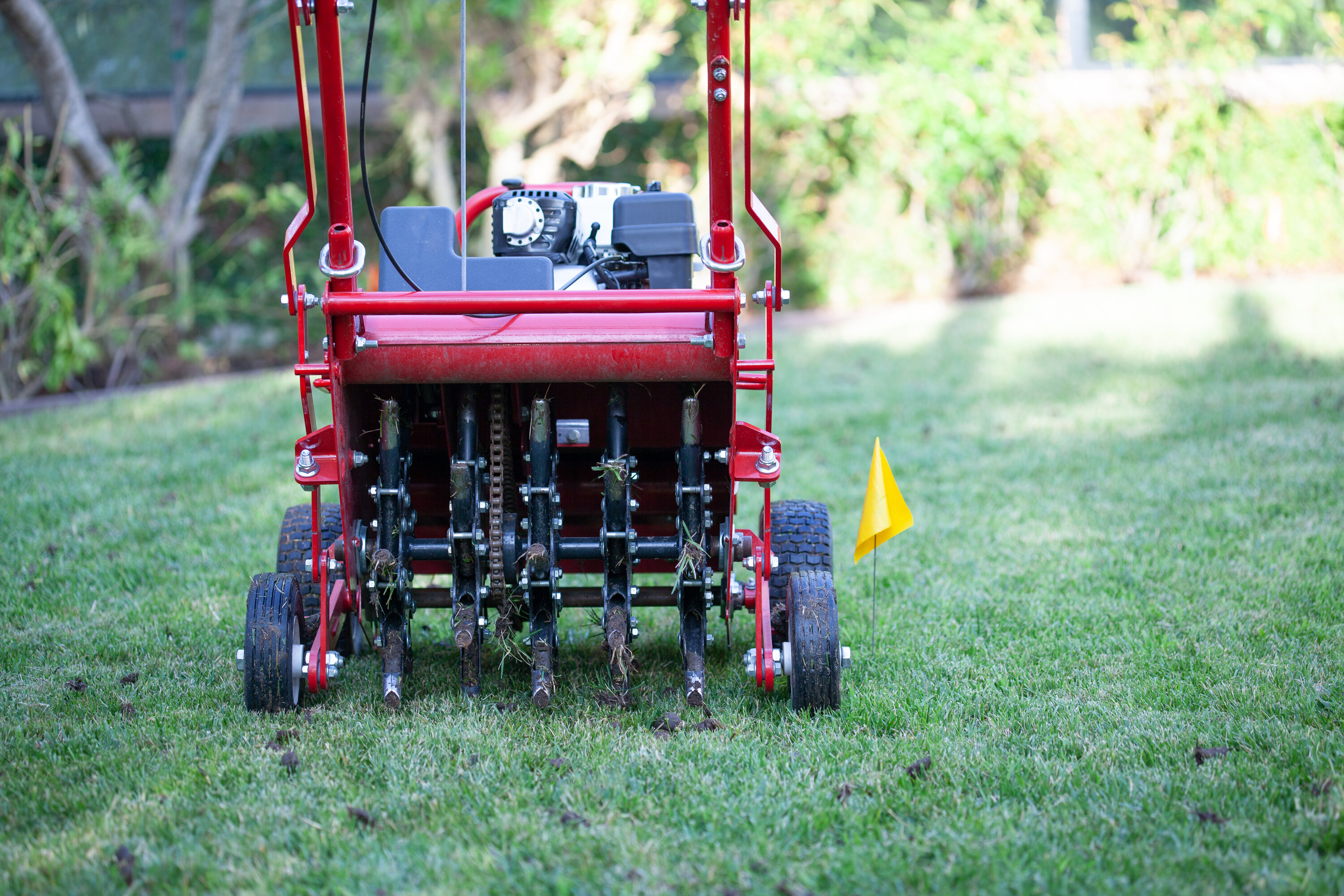 A photo of a red and black lawn aeration machine, sitting on a grass lawn, with a yellow flag next to it