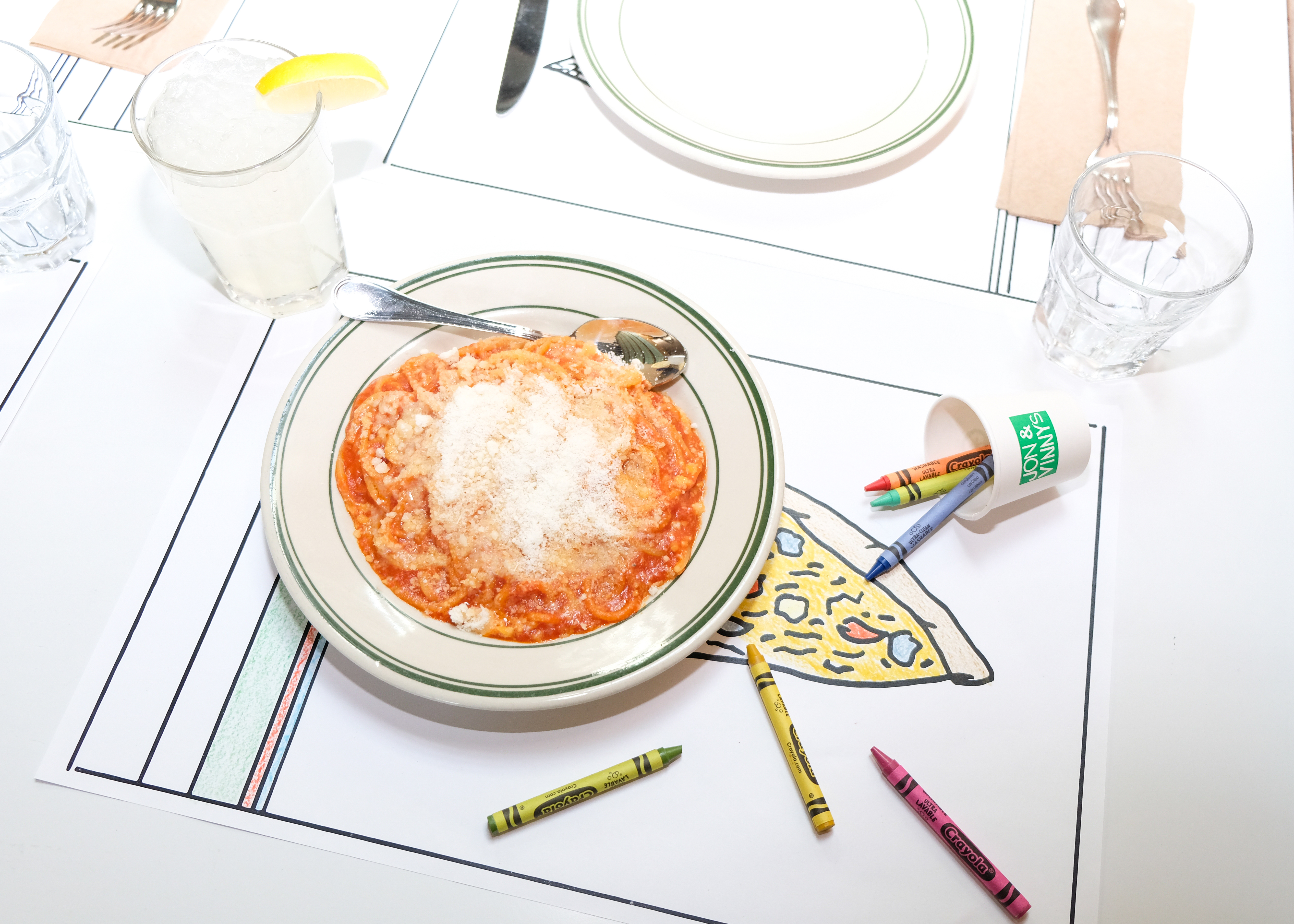 A white paper place mat with a black outline drawing of pizza is colored in with crayons on the table; a bowl of red sauce pasta in a white dish is also on the table.