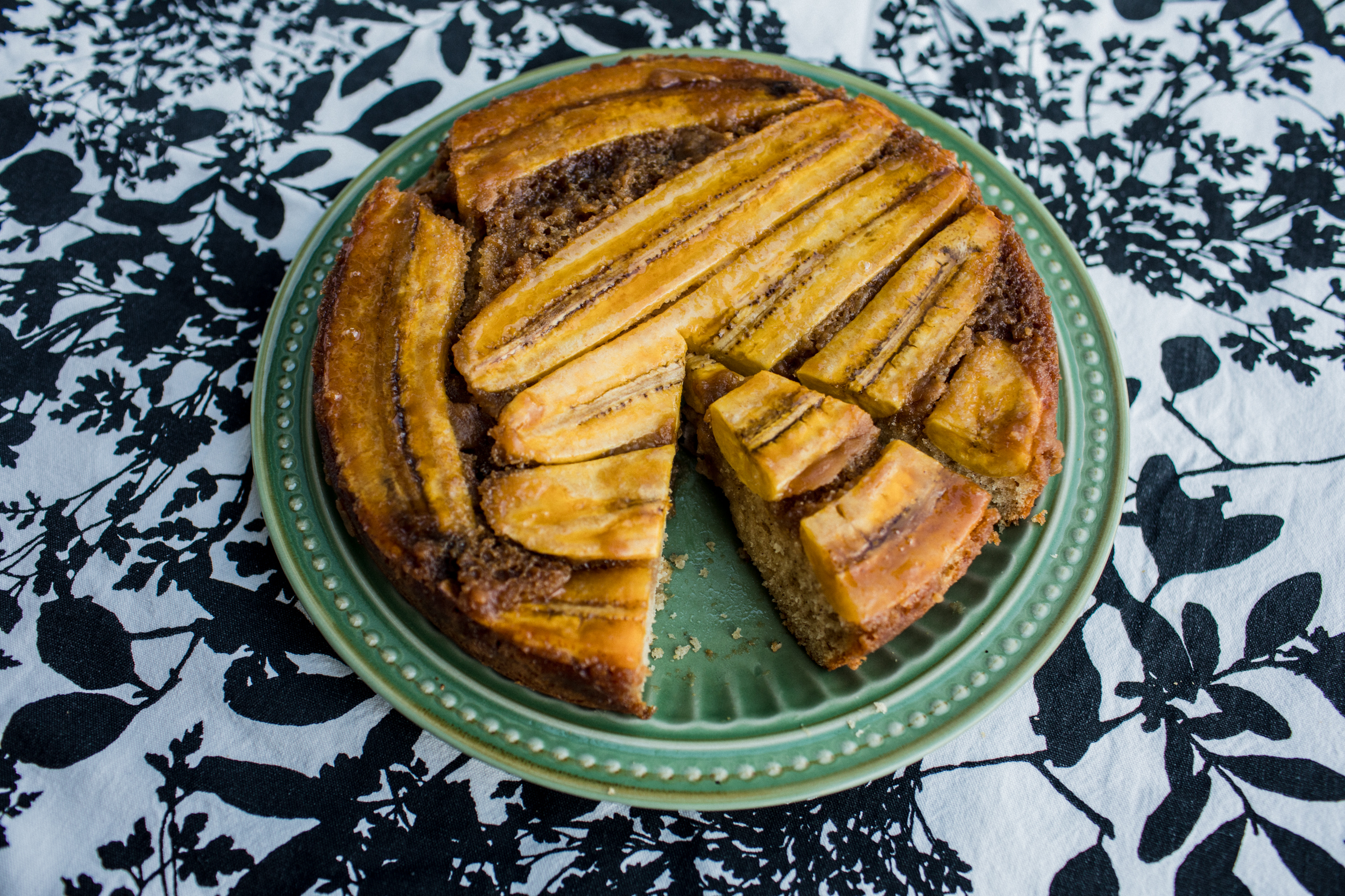 Plantain upside down cake with a slice cut out, served on a green plate on a patterned tablecloth.
