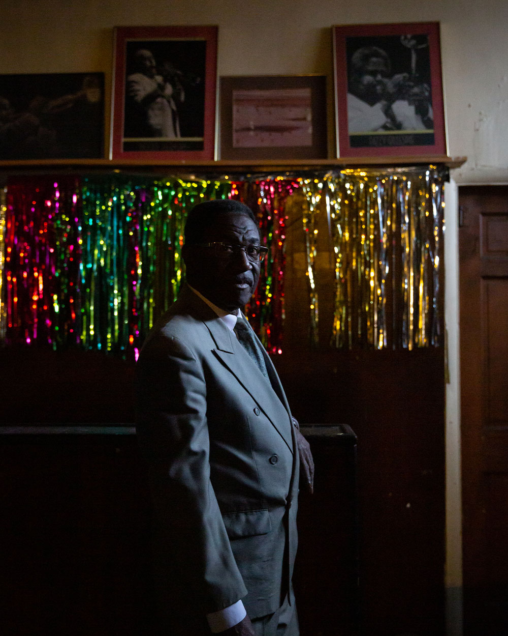 A man in a grey suit stands below photos and tinsel decor at a bar. 