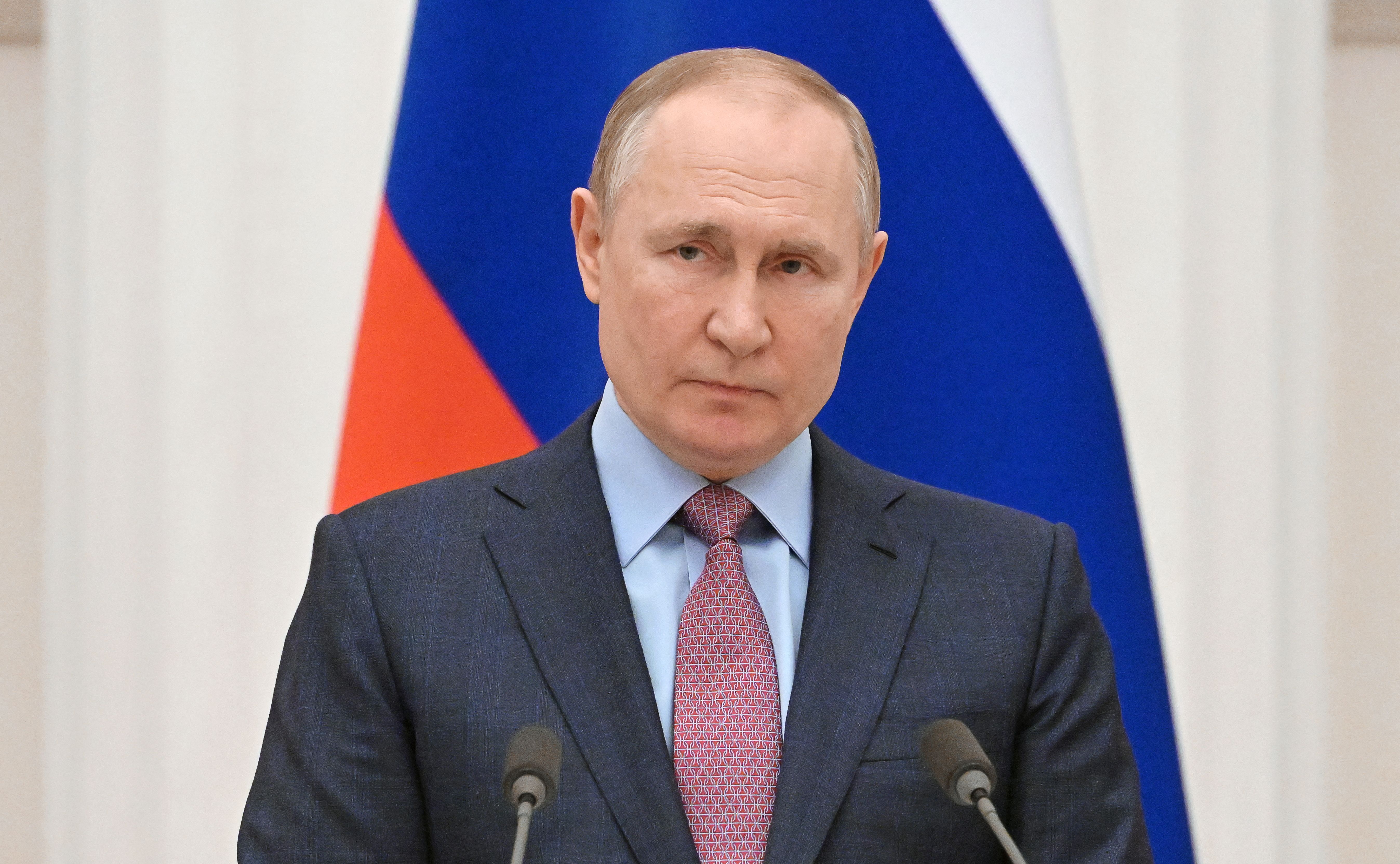 Russian president Vladimir Putin, wearing a suit and tie, stands at a podium in front of a Russian flag.