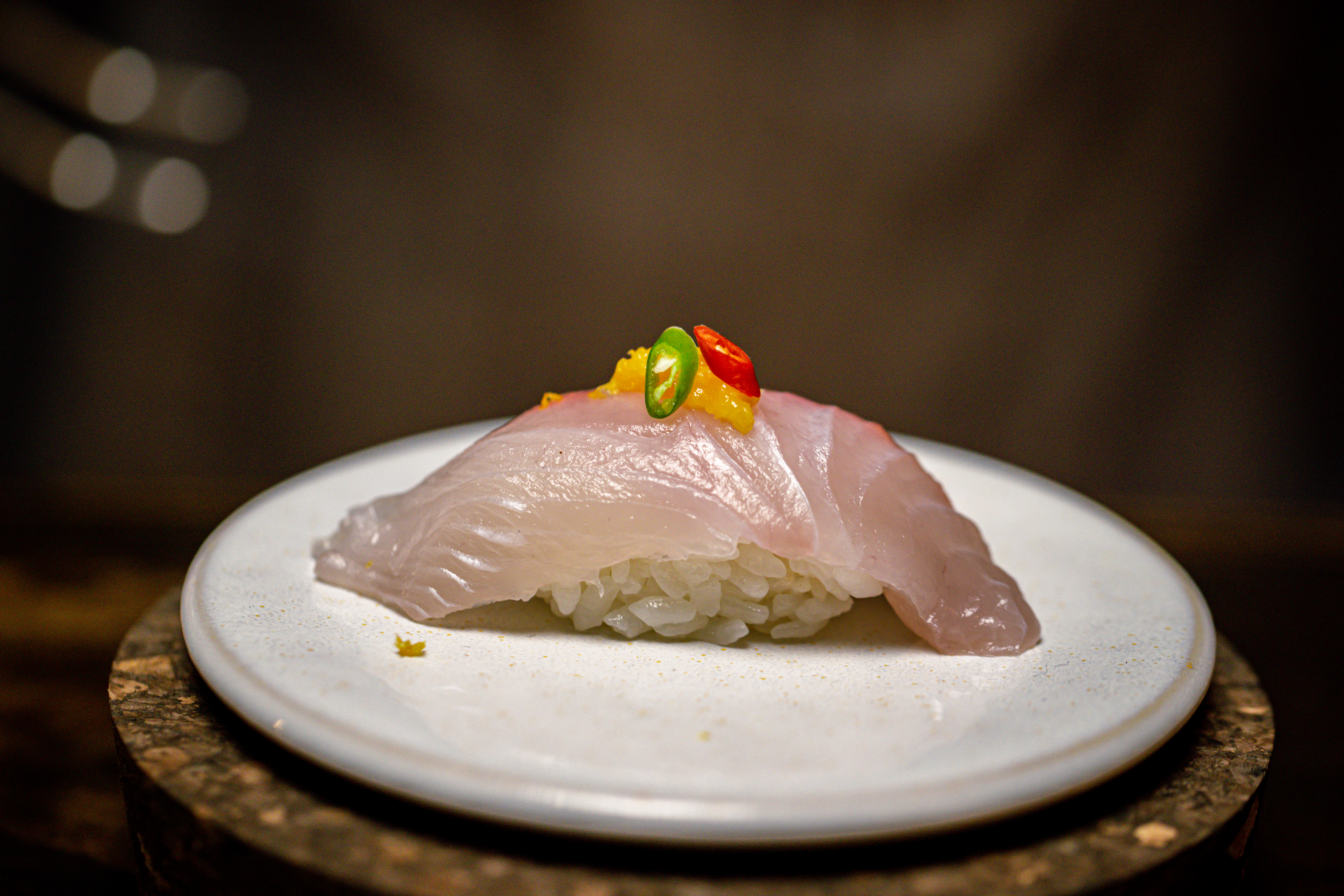 A piece of sushi with a pale pink fish with sliced green and red chiles on a mound of white rice on a round white plate.
