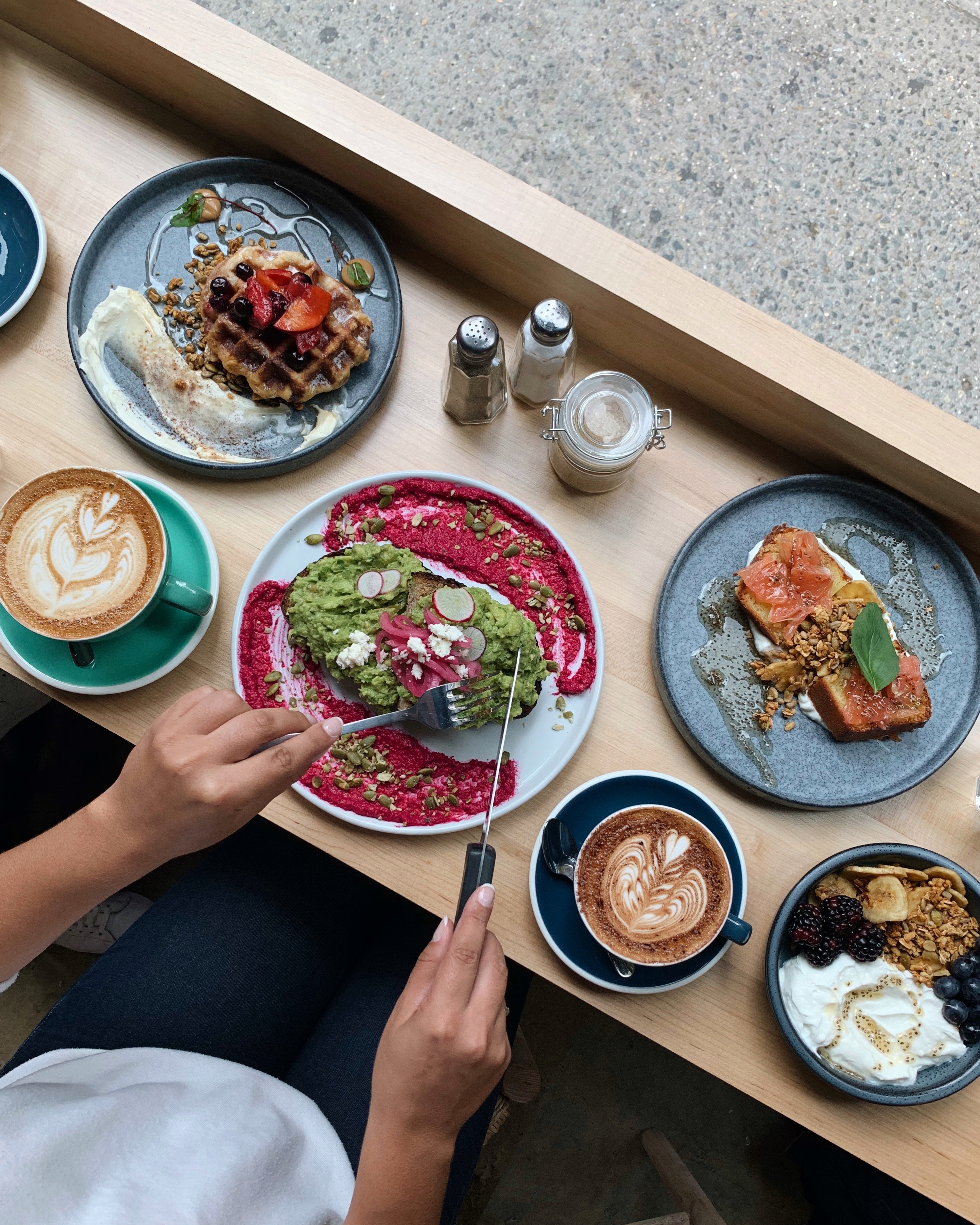 A person slices into an avocado toast plated with a beet puree, a side of waffles, a latte, brekkie bowls, and more.