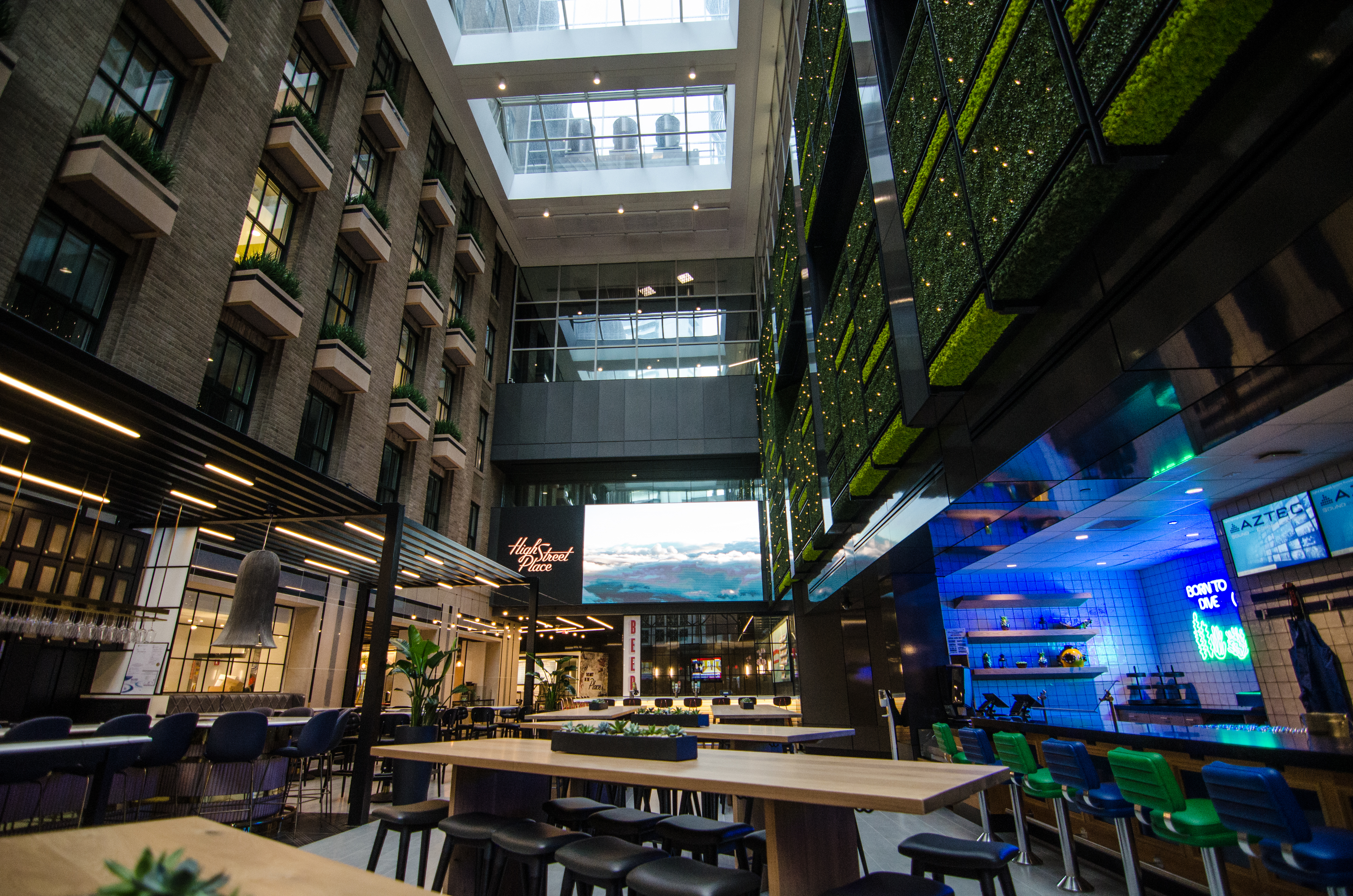 An interior view of a three-and-a-half story atrium with communal seating and a variety of food and beverage vendors. There’s also a green wall and a large video screen.