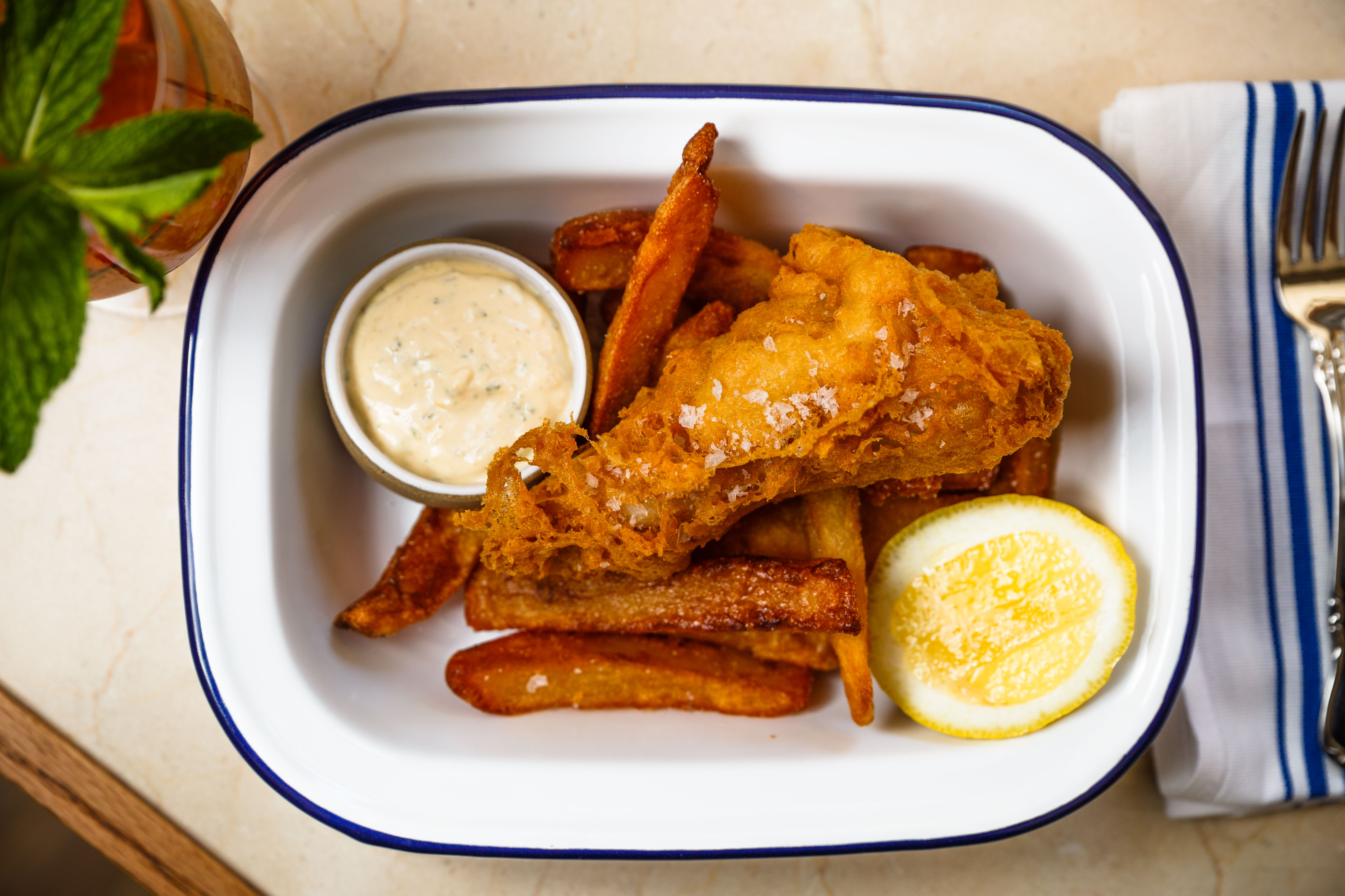 An overhead view of Dame’s fish and chips, featuring a large serving of fried hake, french fries, a ramekin of tartar sauce, and a lemon wedge.