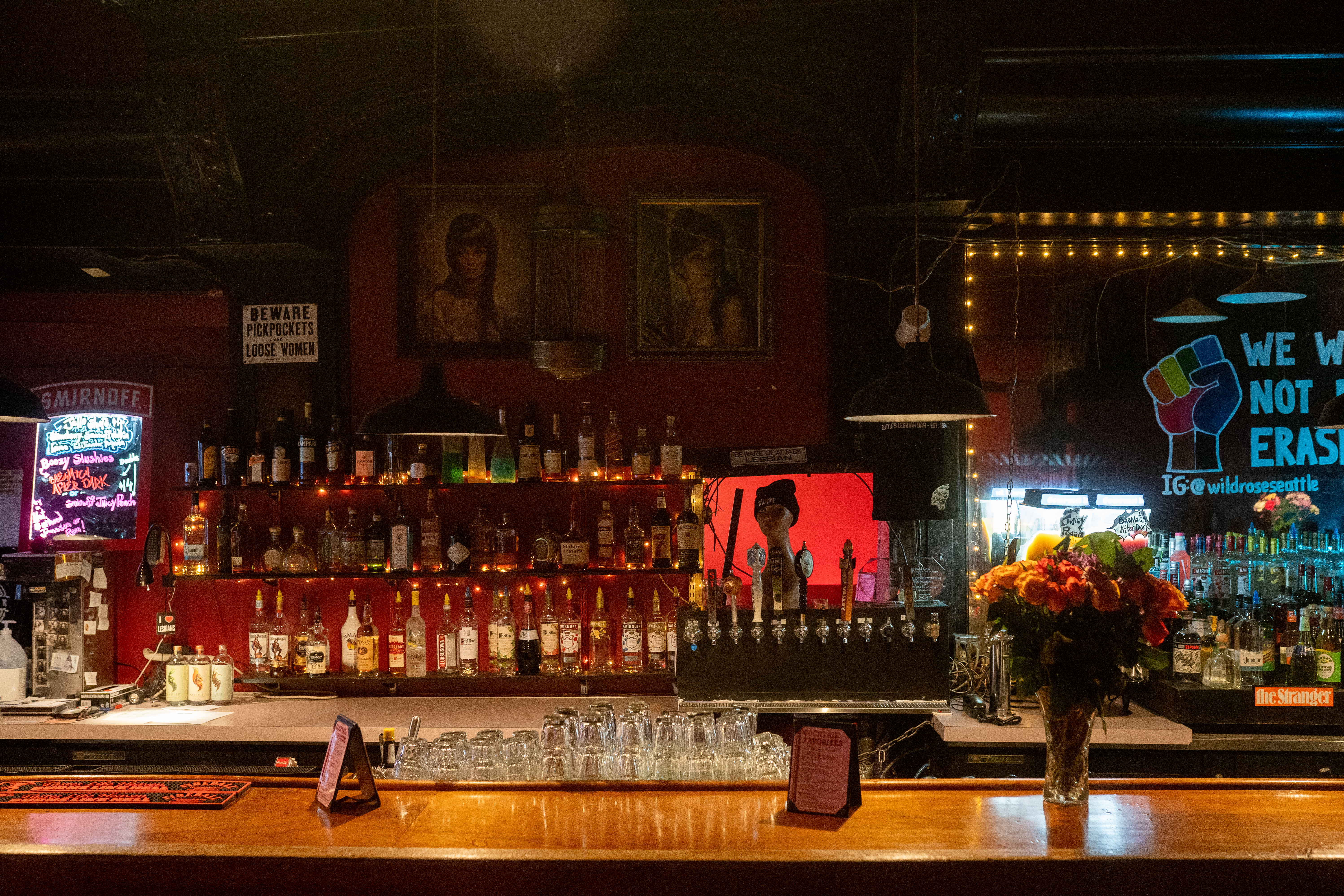 A bar with bottles on racks behind it and signs reading “Beware: pickpockets and loose women” and a sign with a raised rainbow-colored fist with the words “we will not be erased.”
