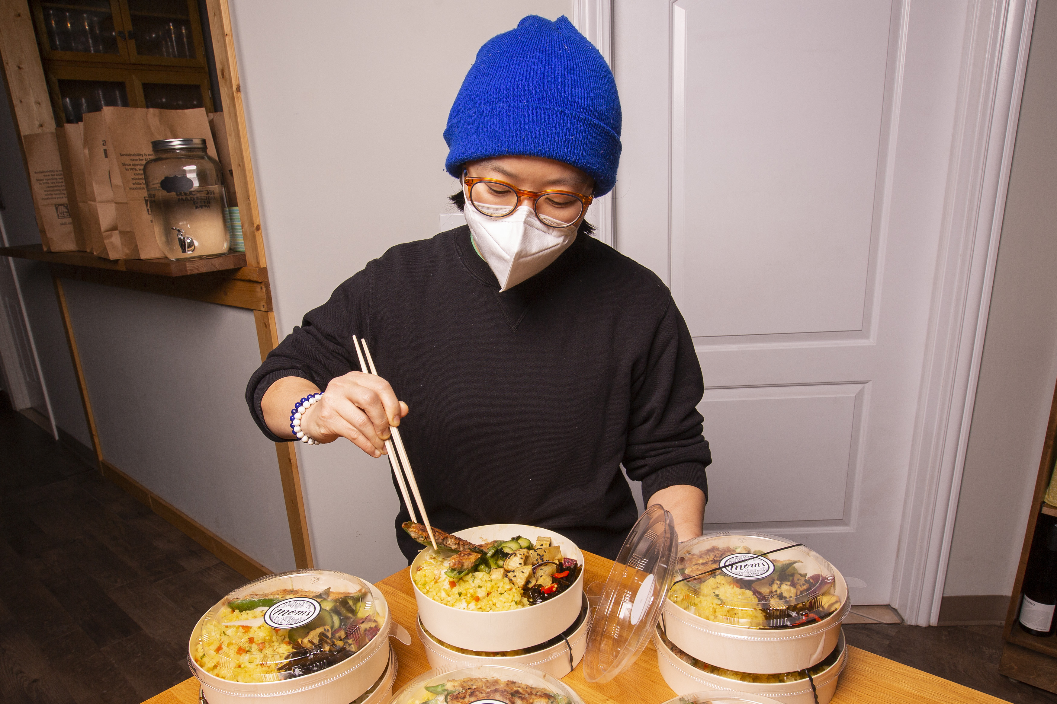 A chef wearing a white mask and blue beanie hat uses chopsticks to arrange food in a round bento box.